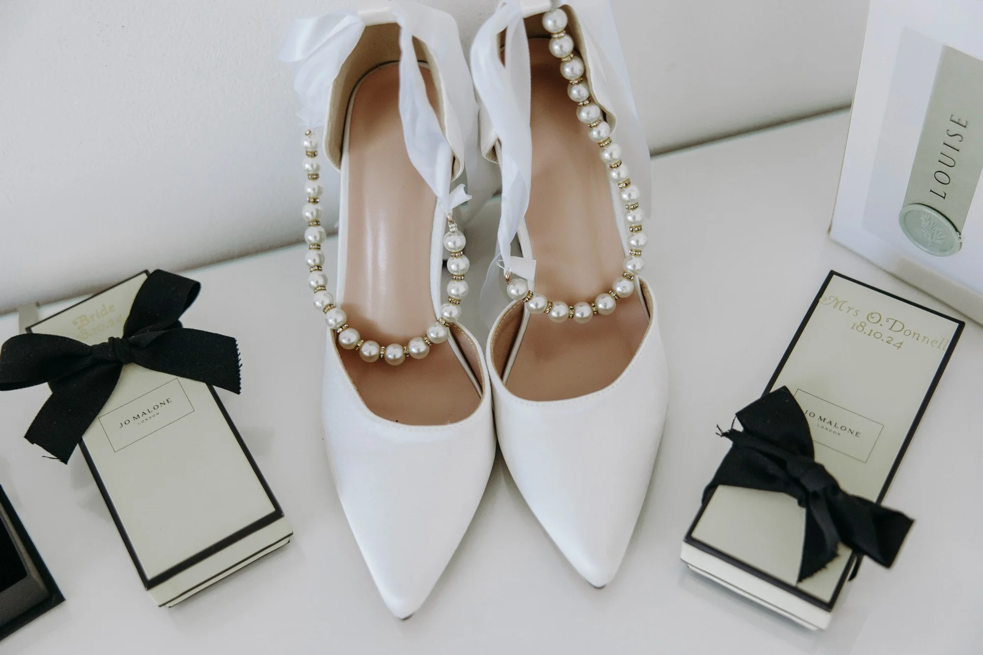 A pair of white high-heeled shoes with pearl-embellished ankle straps, surrounded by boxed gifts with black ribbons, and a framed photograph of a perfume bottle.