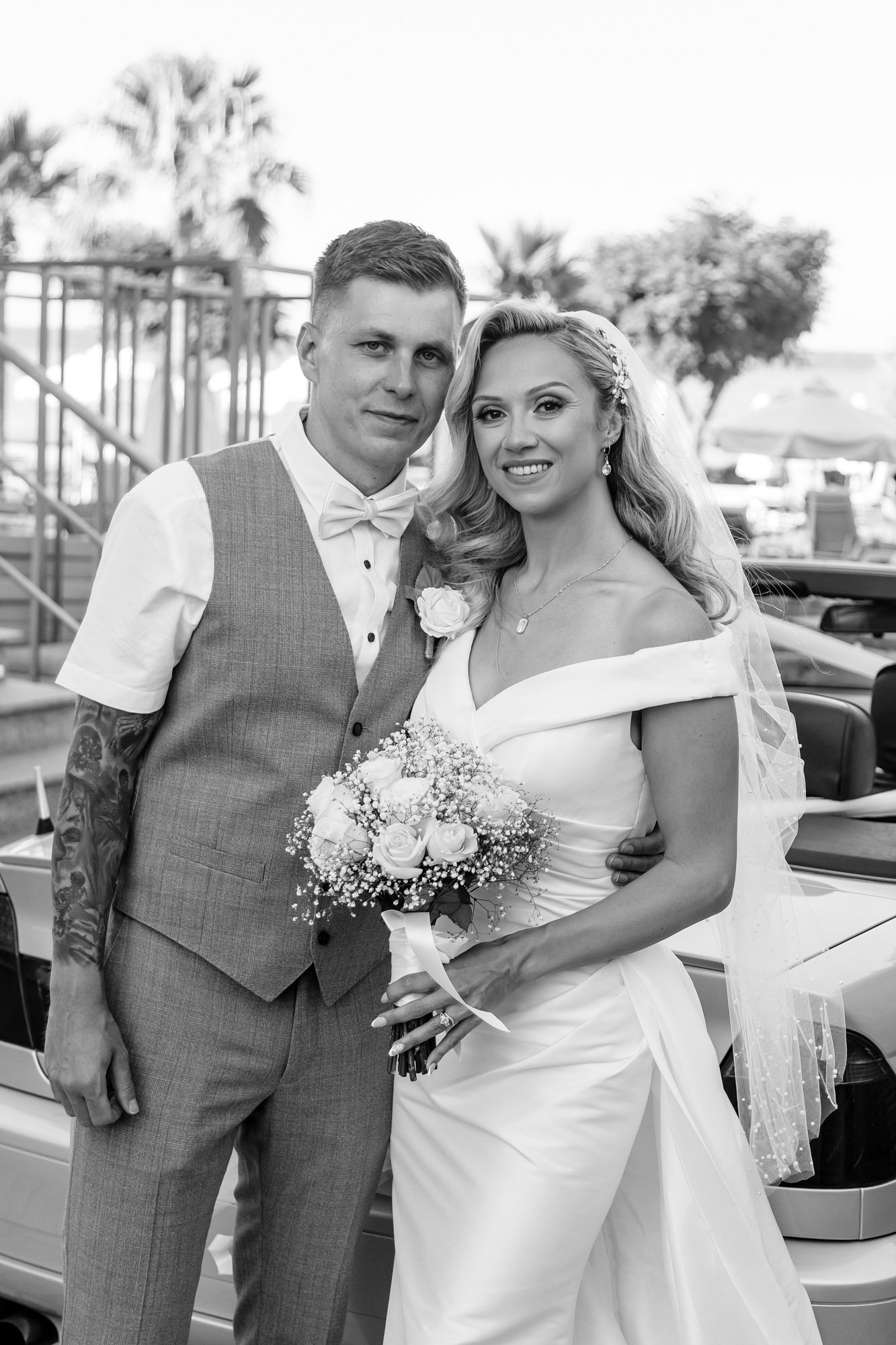Black and white photo of a bride and groom standing together outdoors, smiling, with the bride holding a bouquet of flowers.