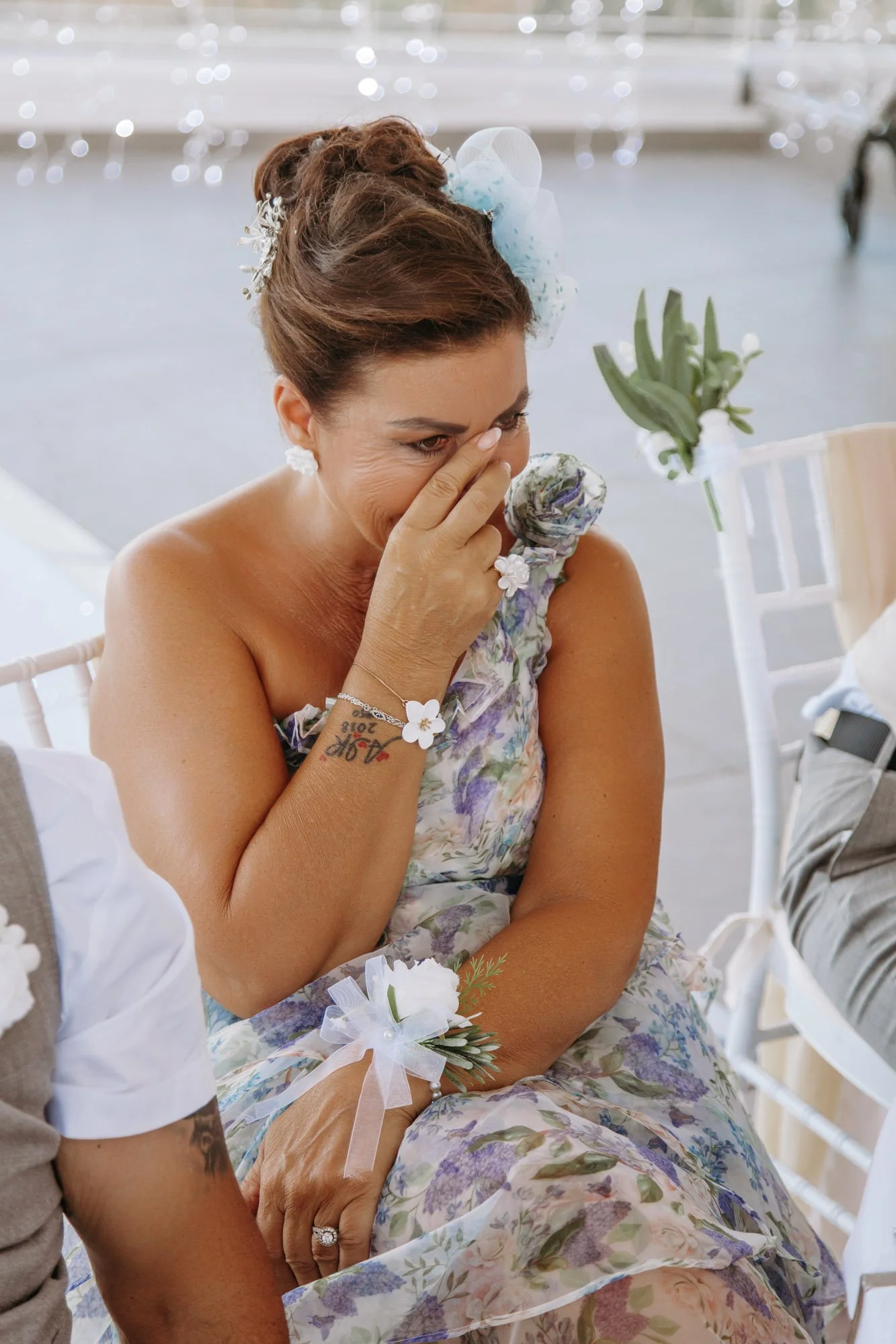 A woman with a floral dress and a corsage on her wrist is sitting and crying, covering her mouth with her hand. She has an emotional expression. The background has blurred lights and white chairs.
