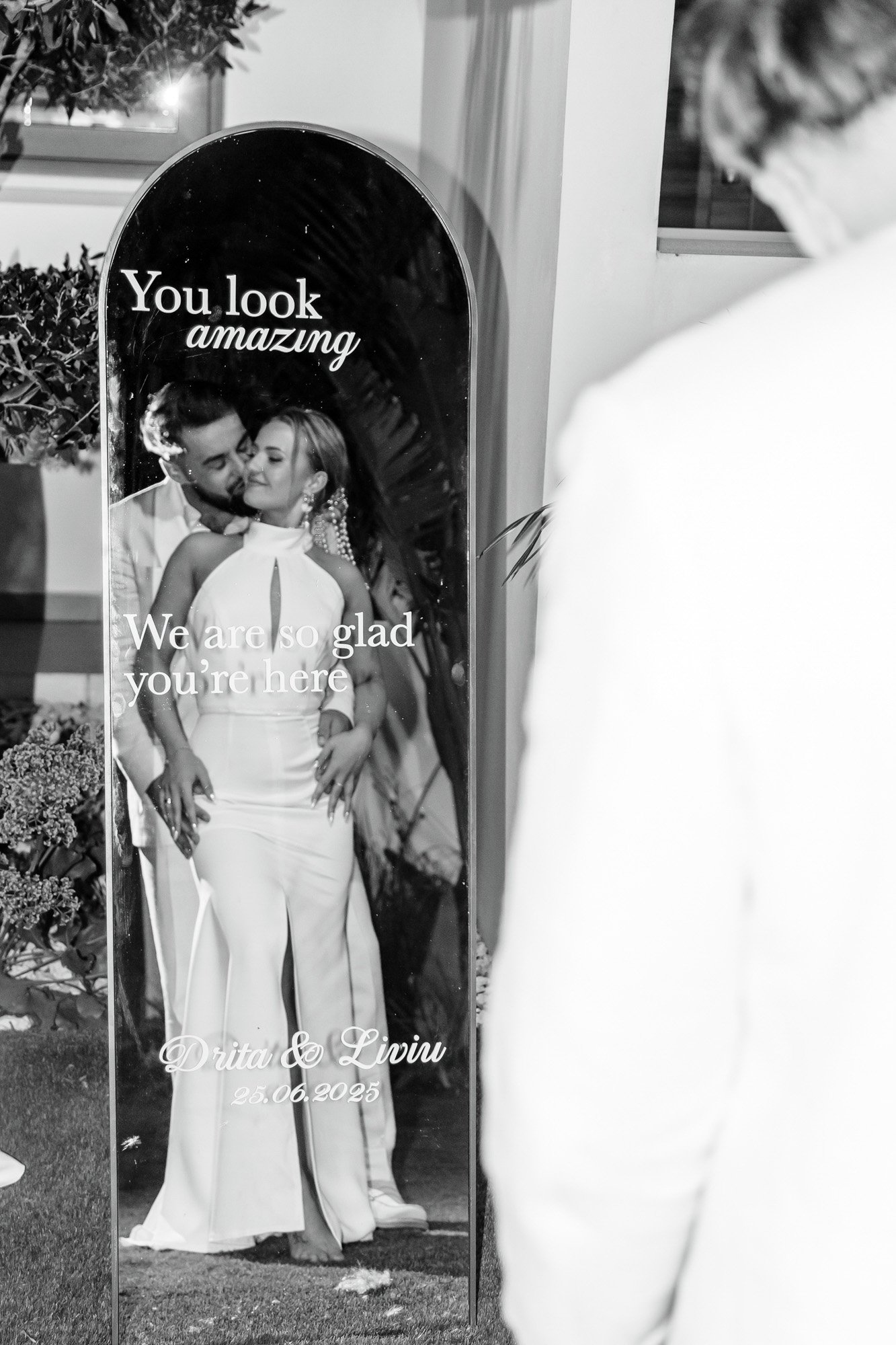 A man and woman embracing in front of a mirror at a wedding, with the words 'You look amazing' and 'We are so glad you're here' written on it. The couple's names, Drita and Liviu, and the date 25.06.2025 are also displayed.