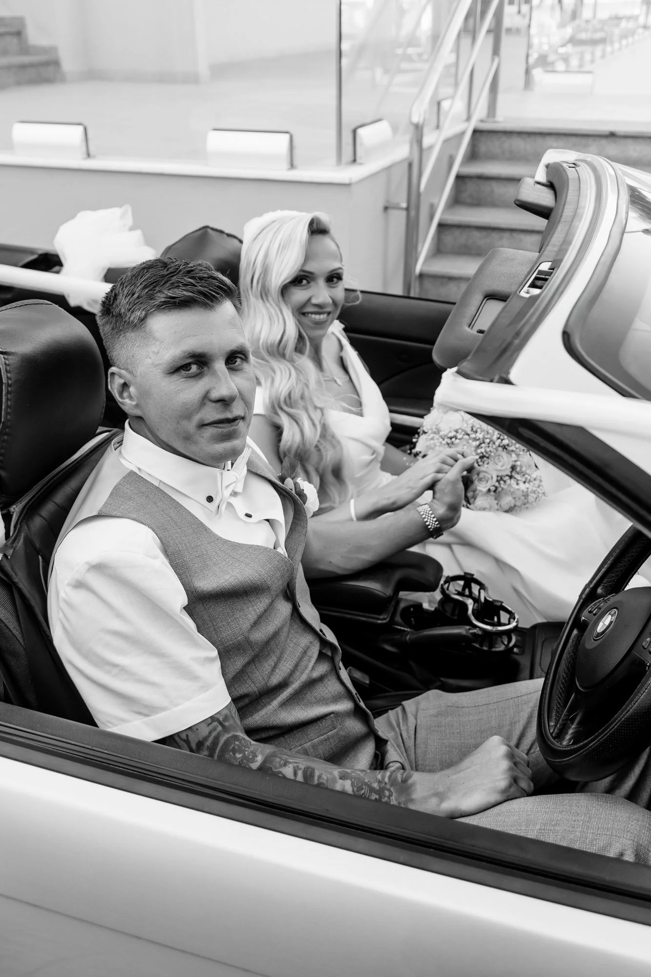 A black and white photo of a bride and groom sitting in a convertible car, with the bride holding a bouquet and smiling, while the groom looks towards the camera.