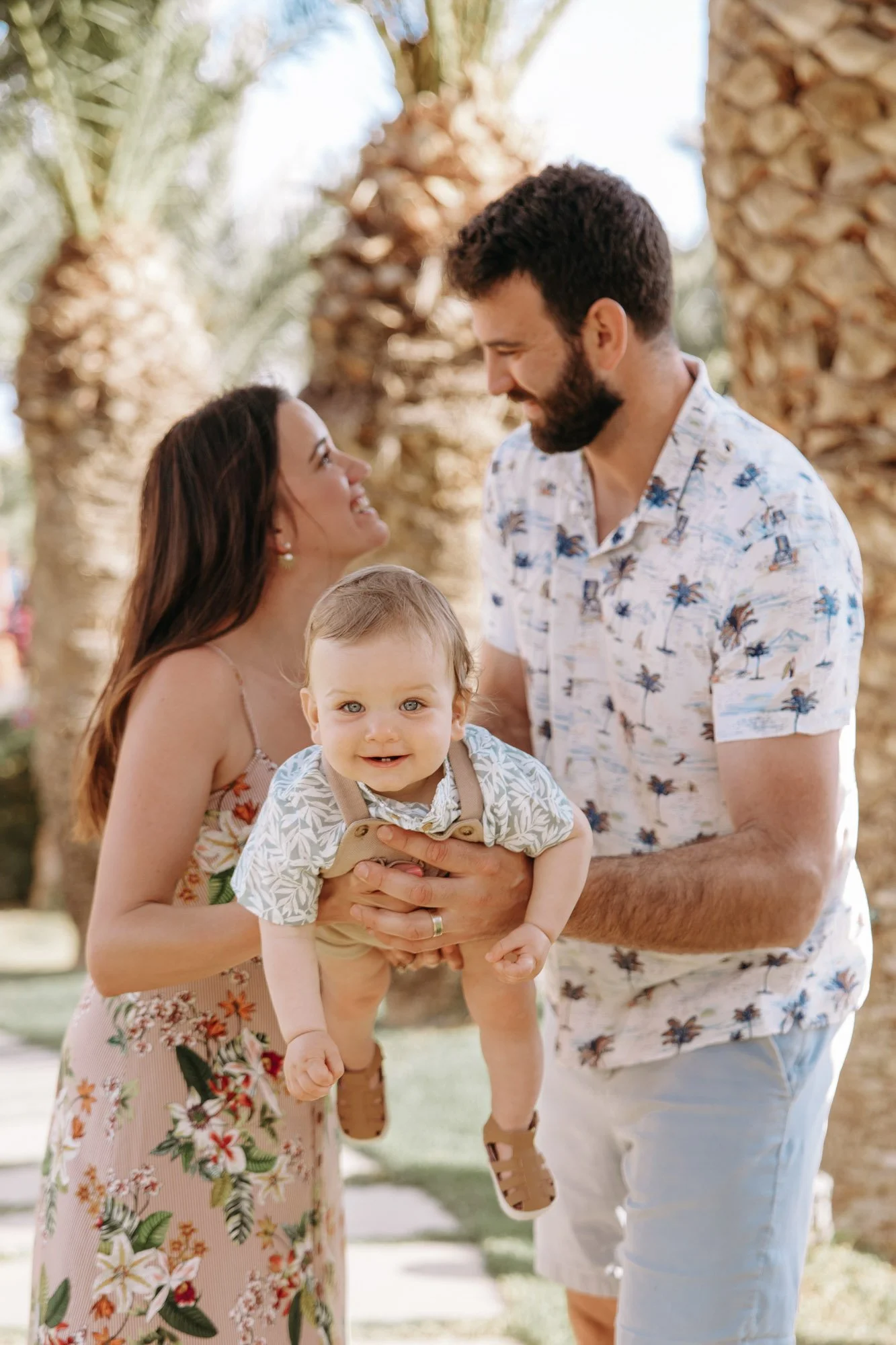 A smiling woman holding a small boy outdoors with a man standing nearby, with palm trees in the background.
