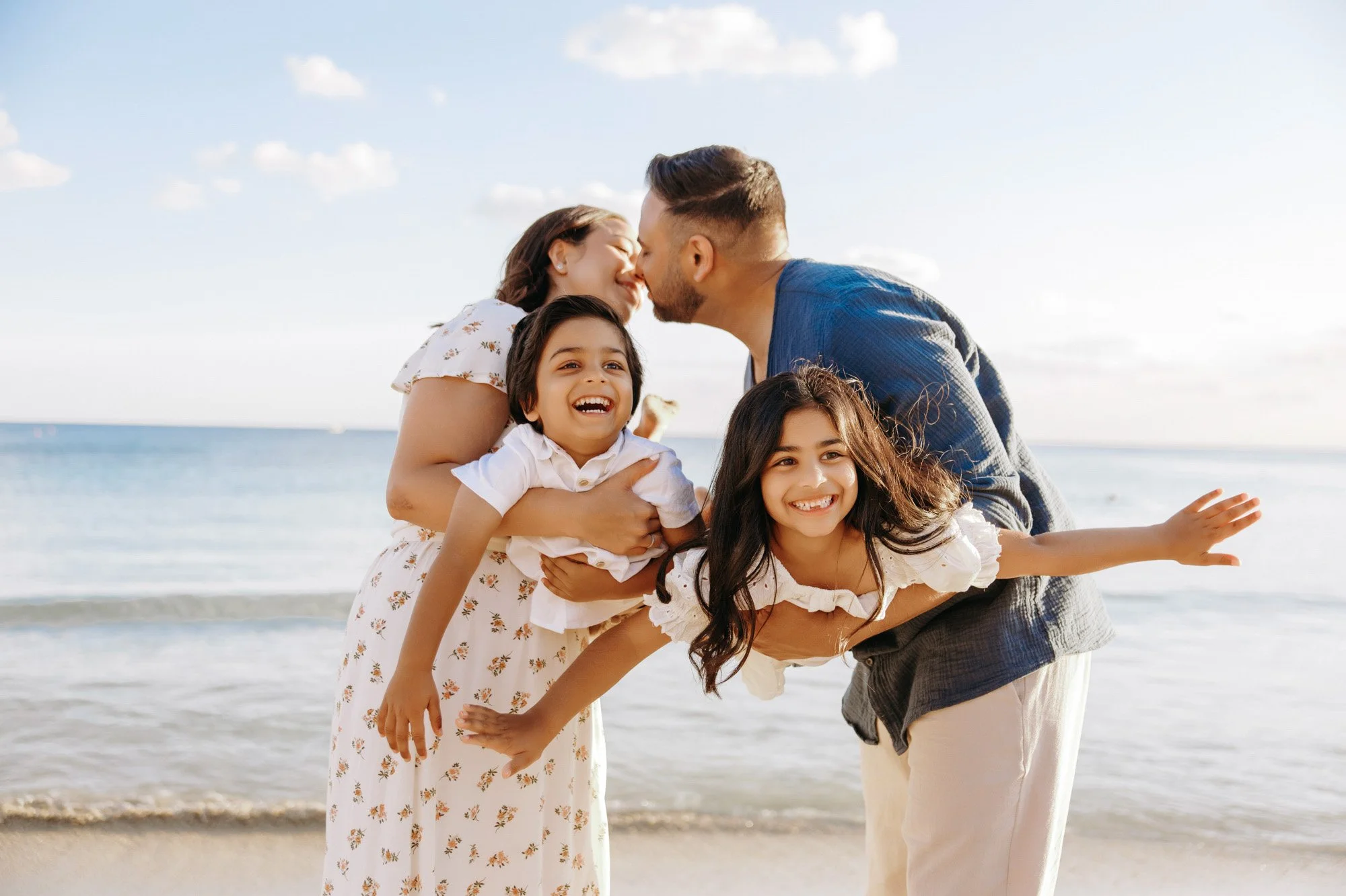 A family of four on the beach, with two young girls and their parents, sharing a joyful moment with the mother and father leaning in to kiss, while the daughters smile and pose for the photo.