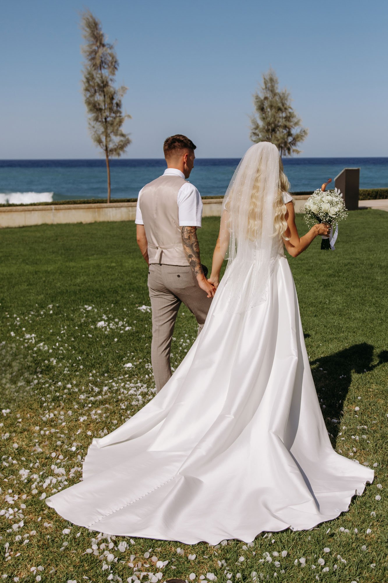 Bride and groom holding hands at the beach, the bride wearing a white wedding dress and veil with a bouquet, the groom in a light beige vest and pants, facing the ocean with a clear blue sky.