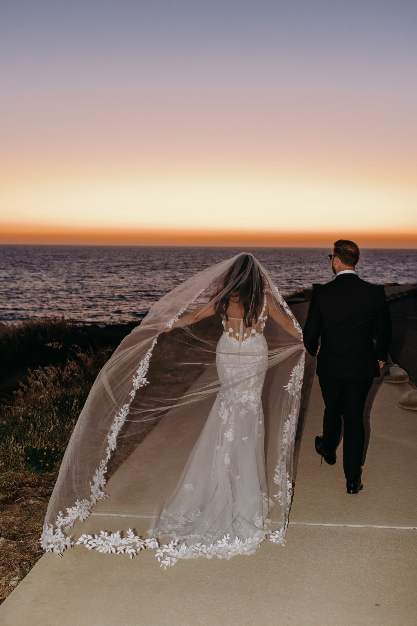 A bride and groom walking along a pathway by the ocean at sunset. The bride is wearing a white lace wedding gown with a long, sheer veil. The groom is dressed in a black suit.