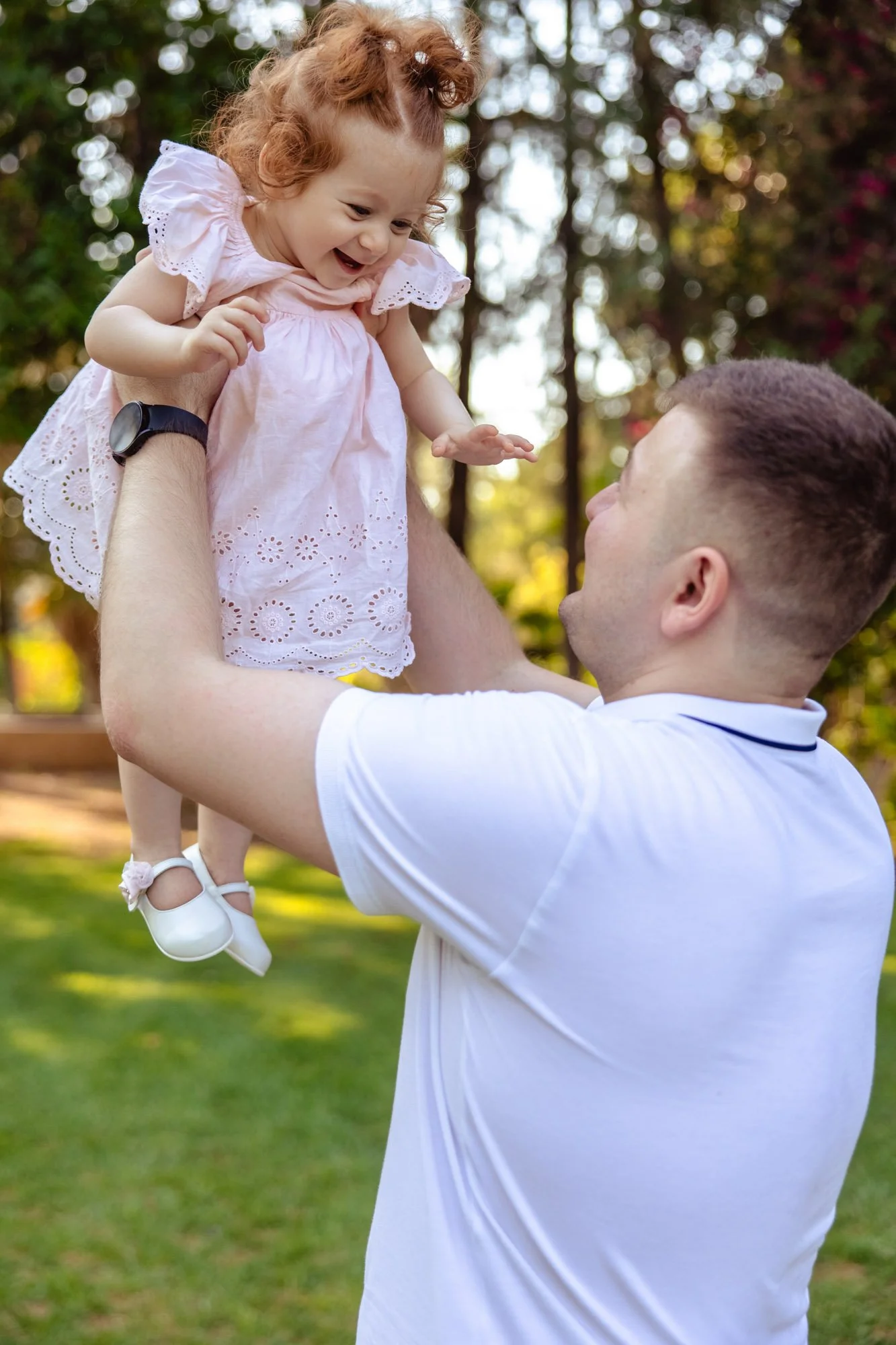 A man lifting a little girl in a pink dress outdoors, smiling and looking at each other with trees in the background.