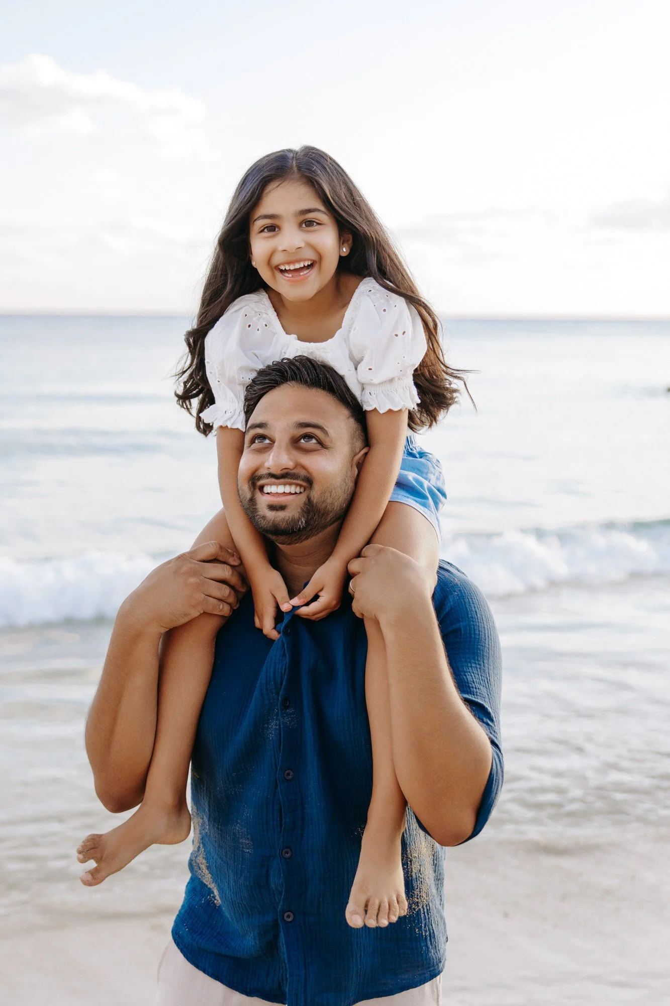 A man giving a girl a piggyback ride on the beach, both smiling, with ocean waves in the background.