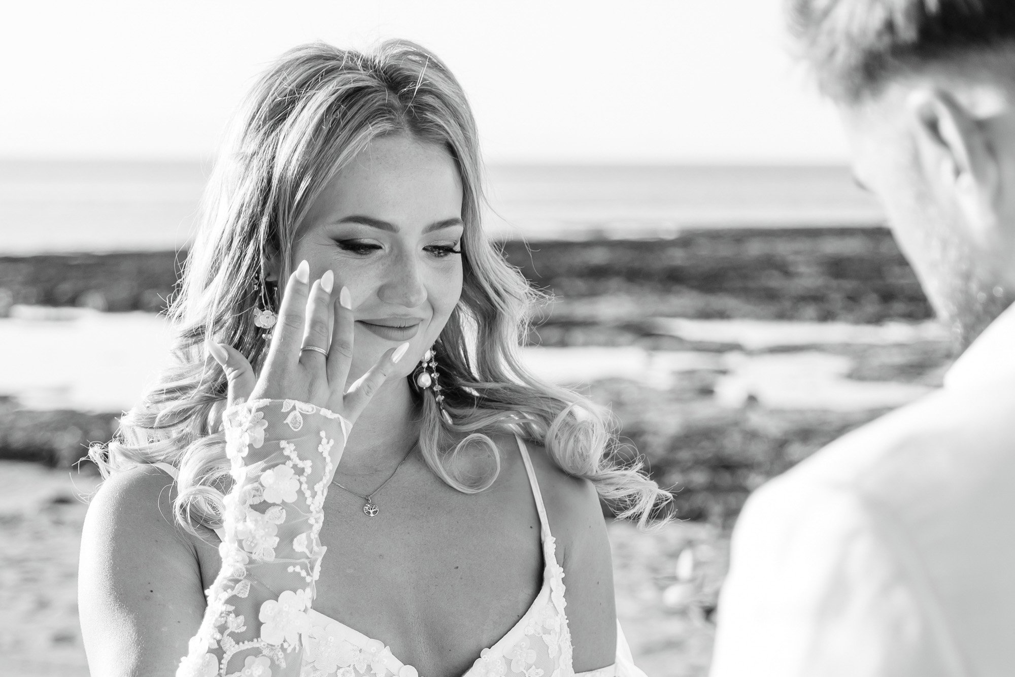 A woman with long wavy hair and floral earrings looks down at her hand, which she is showing to a man. She wears a lace top and a necklace, and her hand is showing a ring. They are standing on a beach.