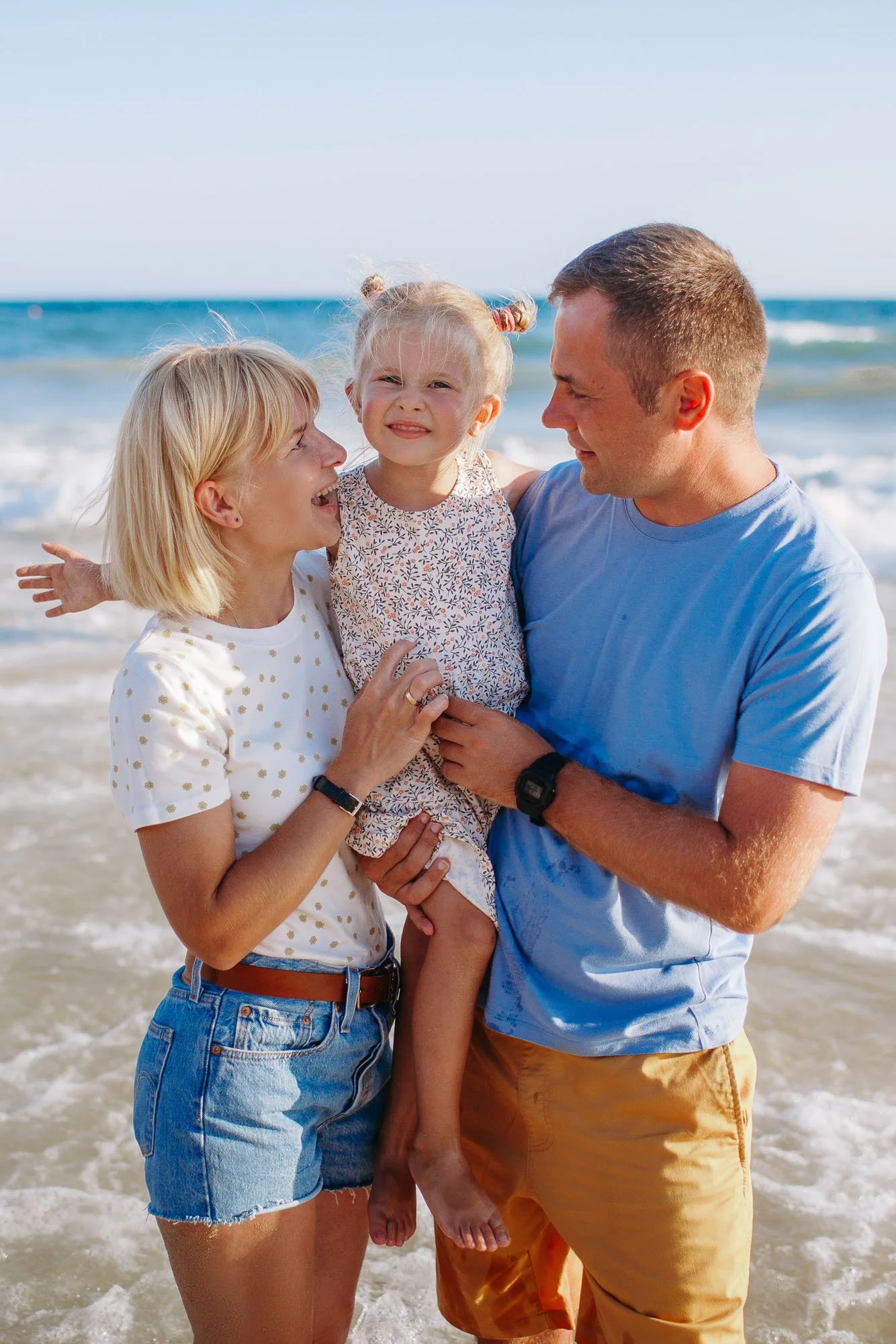 Family of three at the beach, smiling and enjoying a sunny day, with the ocean and waves in the background.