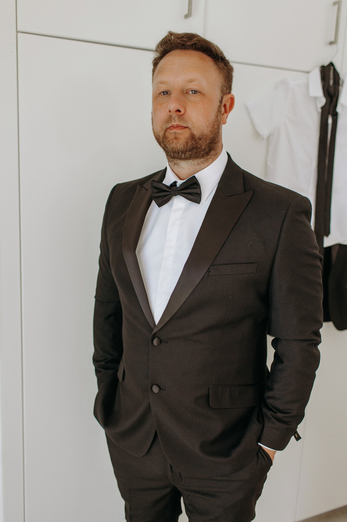 A man in a black tuxedo with a bow tie, standing with his hands in his pockets, in front of a white wardrobe with clothes hanging on the side.