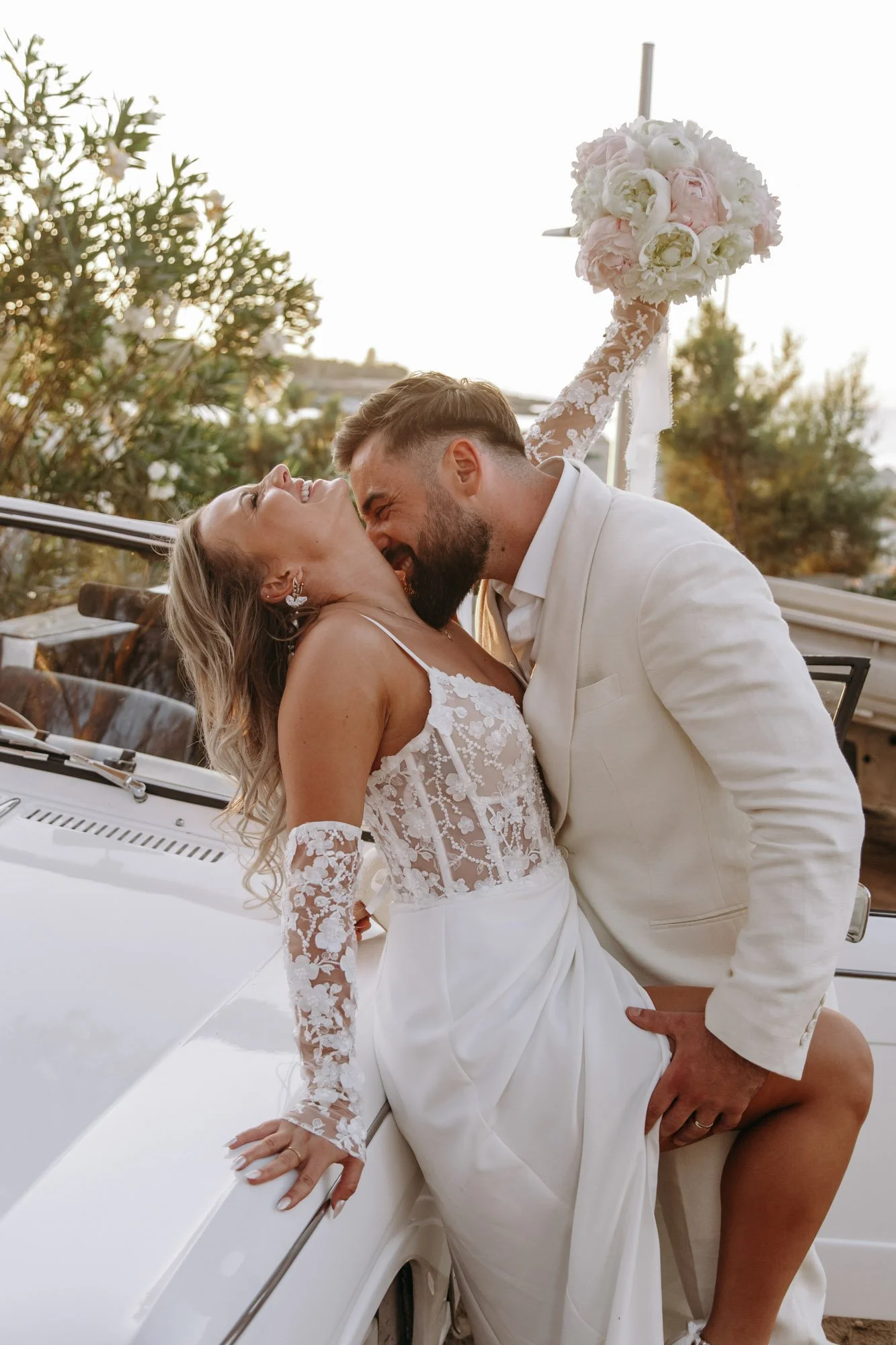 A newlywed couple sharing a joyful kiss outdoors, with a bride in a lace wedding dress and a groom in a white suit, near a classic white car and a backdrop of greenery and a sunset sky.