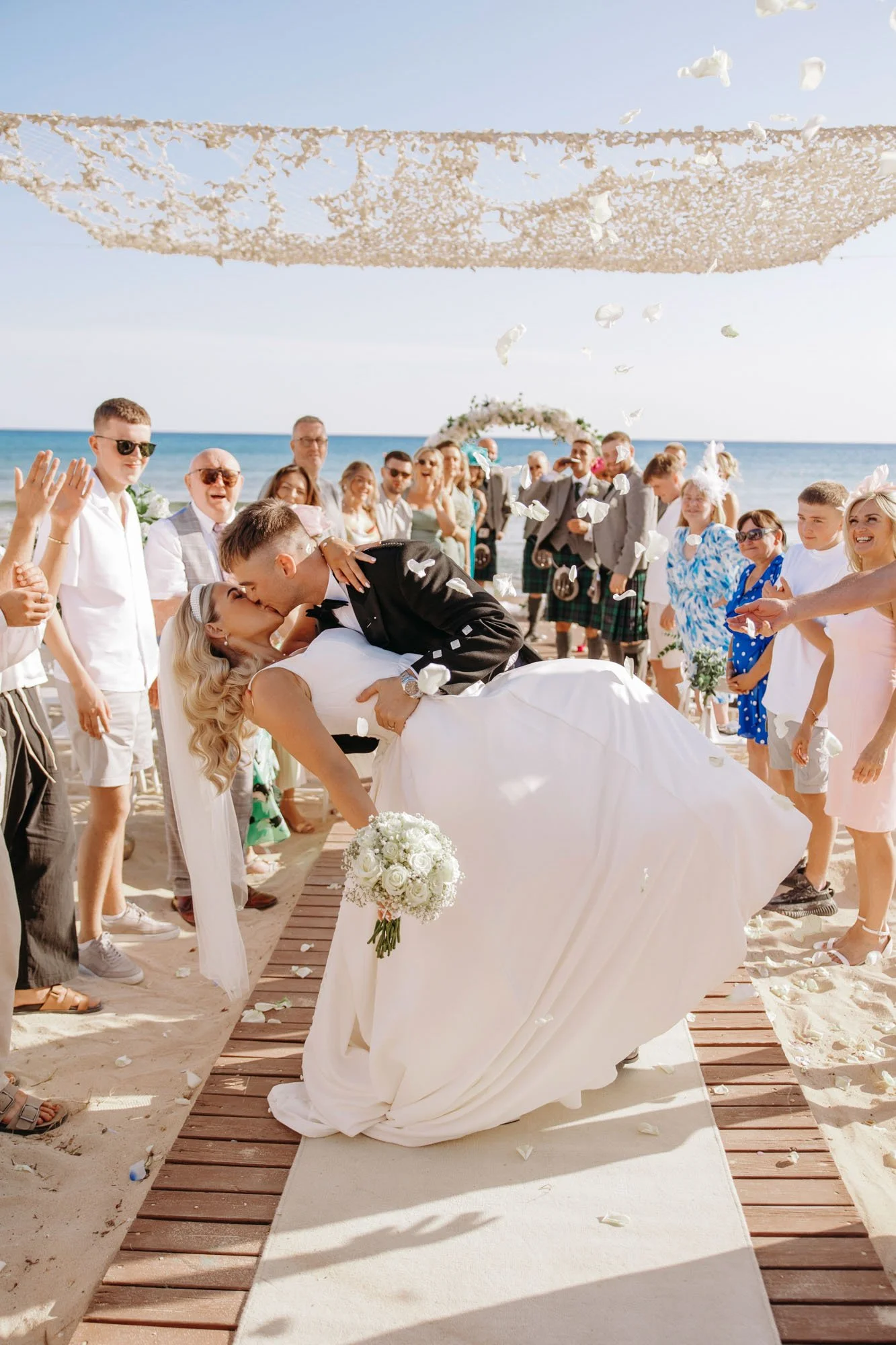 A bride and groom kissing during a beach wedding ceremony with guests celebrating around them and the ocean in the background.
