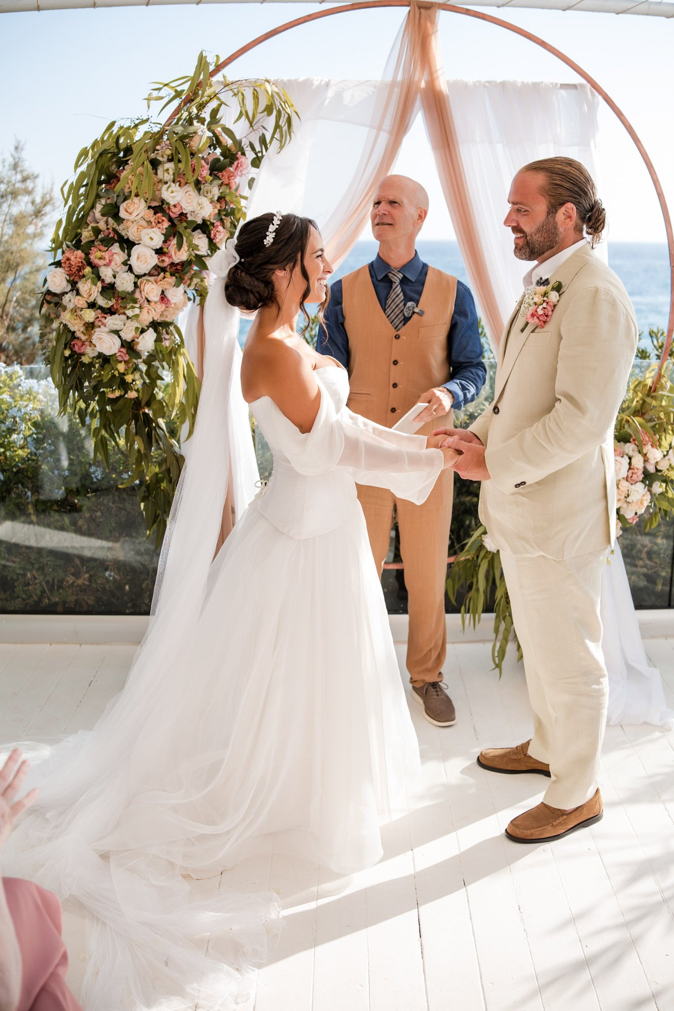 A wedding ceremony by the ocean with a bride and groom holding hands and exchanging vows, under a floral arch with pink and white flowers, officiated by a man in a tan vest, with the sea in the background.