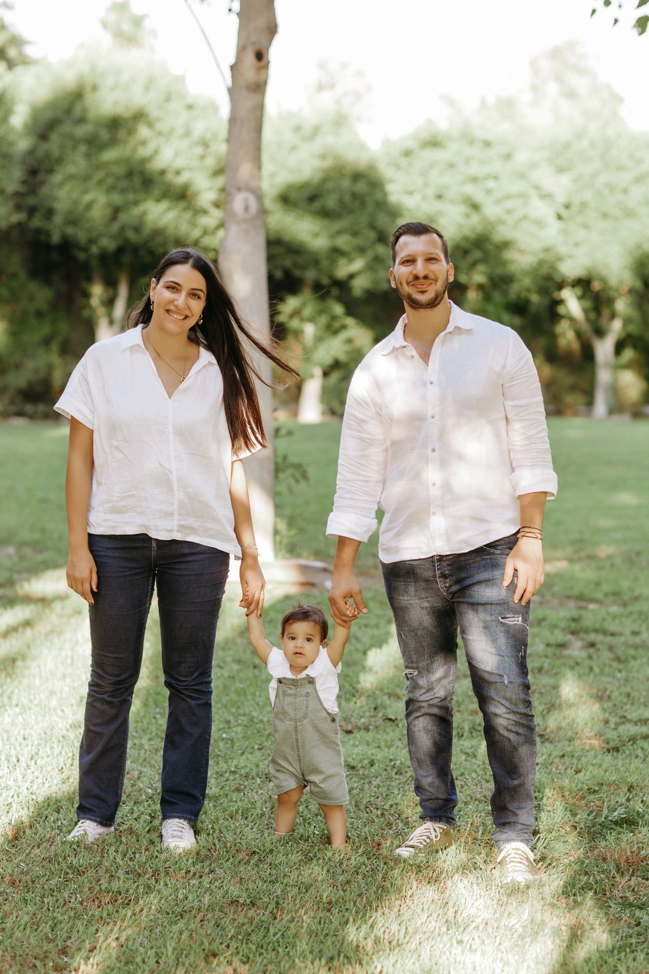 A family of three, two adults and a toddler, walking in a park on a sunny day, holding hands.