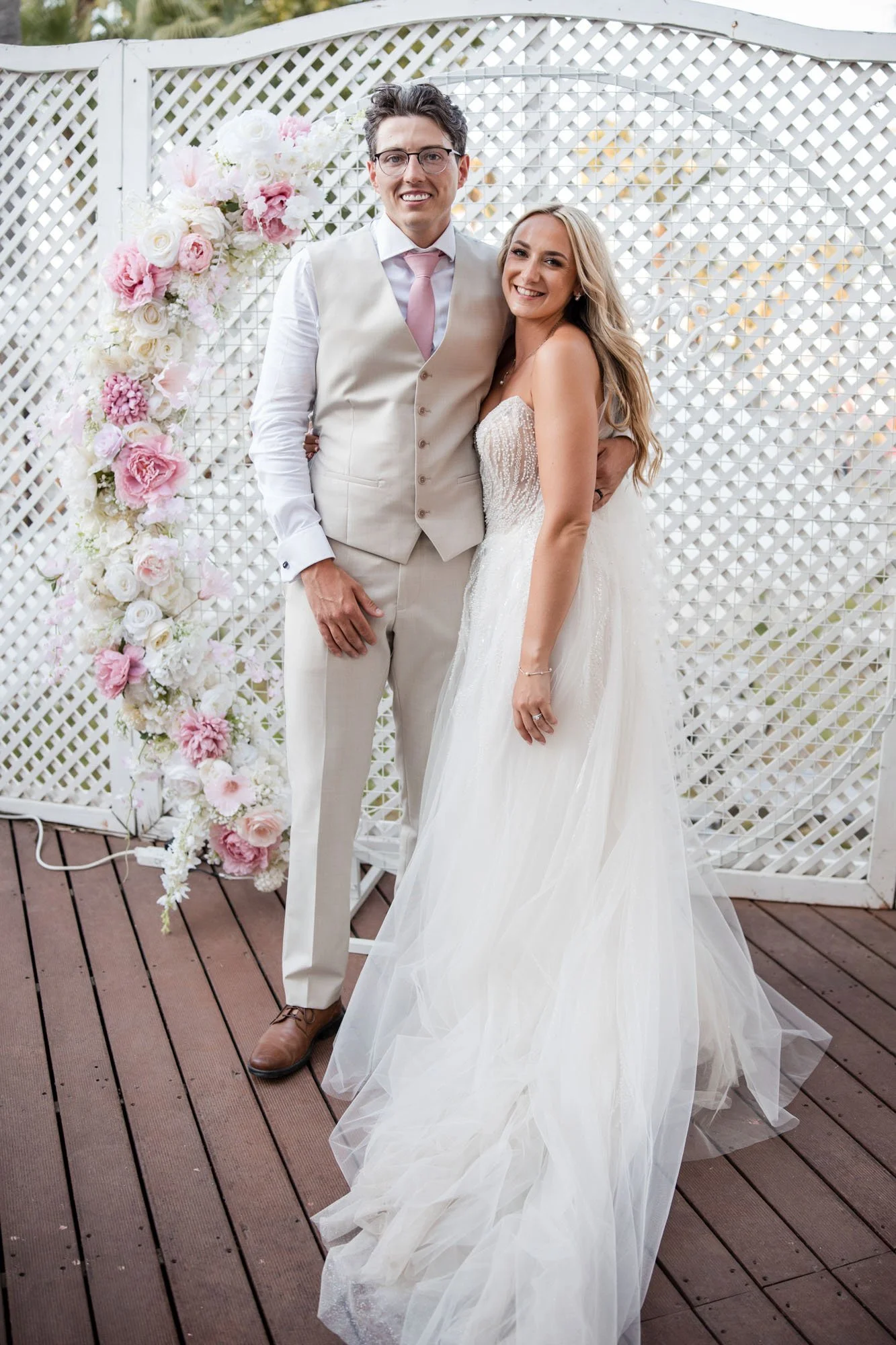 A bride and groom posing together at their wedding on a wooden deck with a lattice backdrop decorated with white and pink flowers.