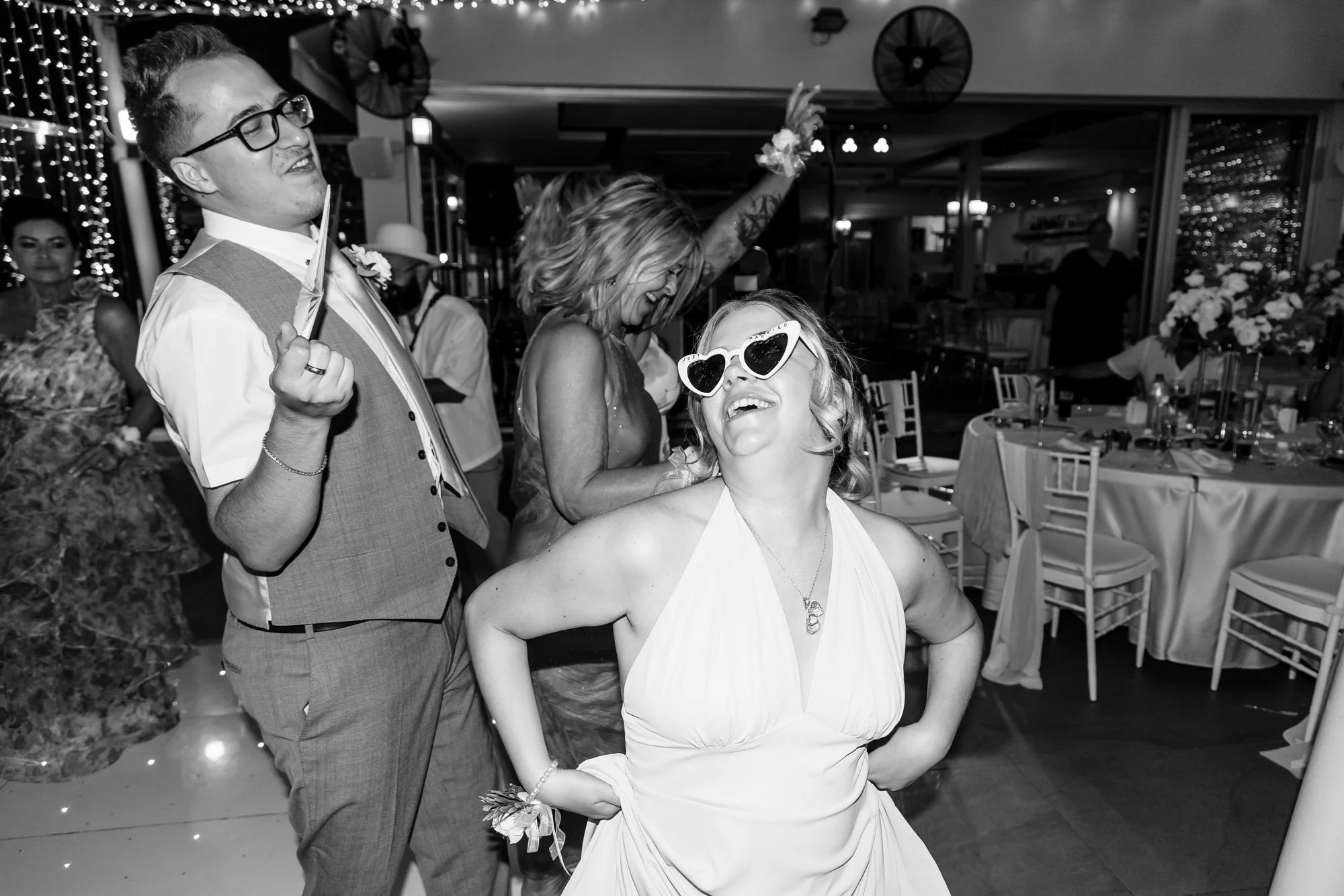 People celebrating at a wedding reception, with a woman wearing sunglasses and a sleeveless dress, and others dancing and enjoying the event.