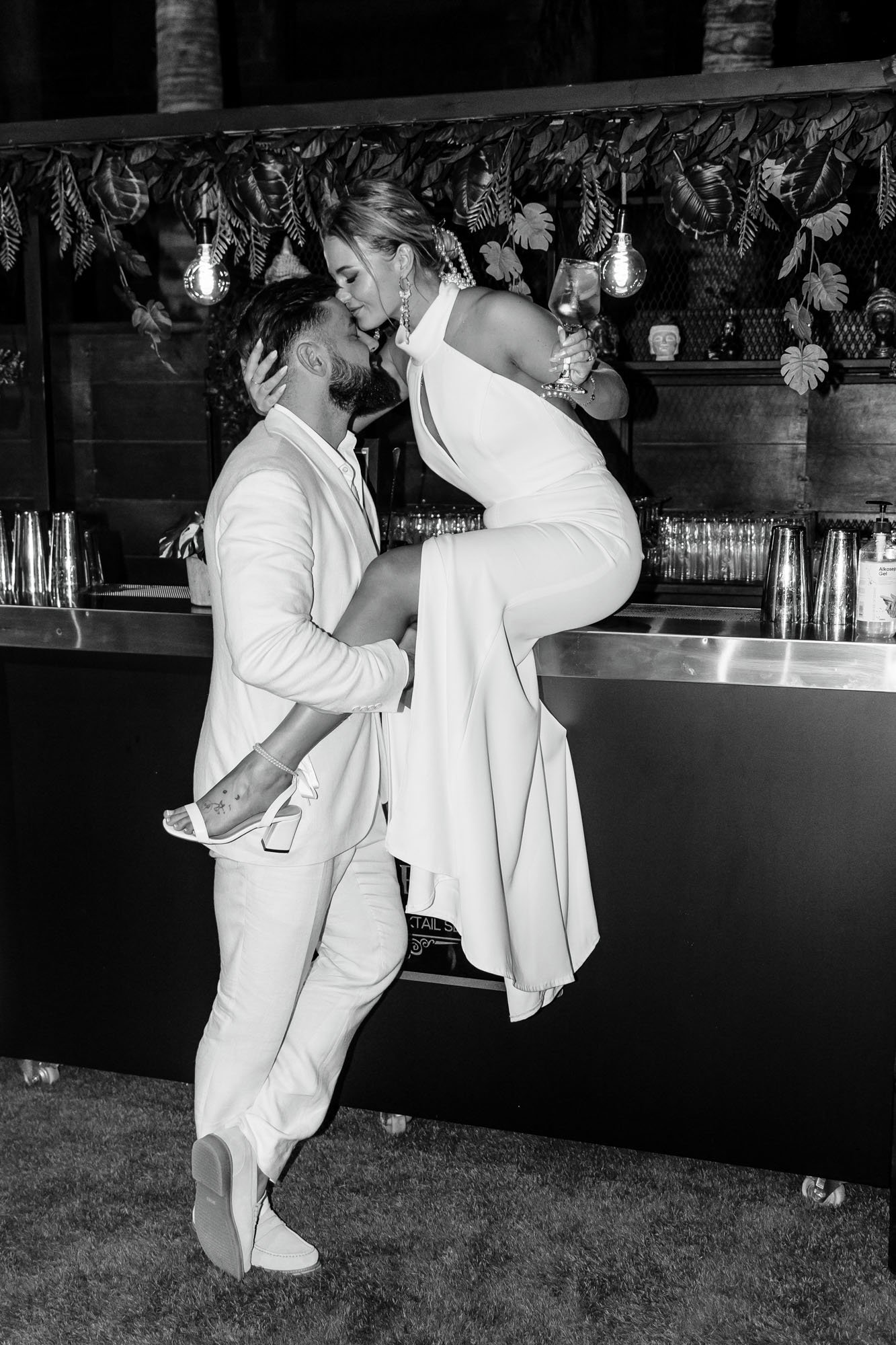A black and white photo of a man in a light suit lifting a woman in a white dress and high heels onto a bar, both leaning in for a kiss, with a decorated background and barware behind them.