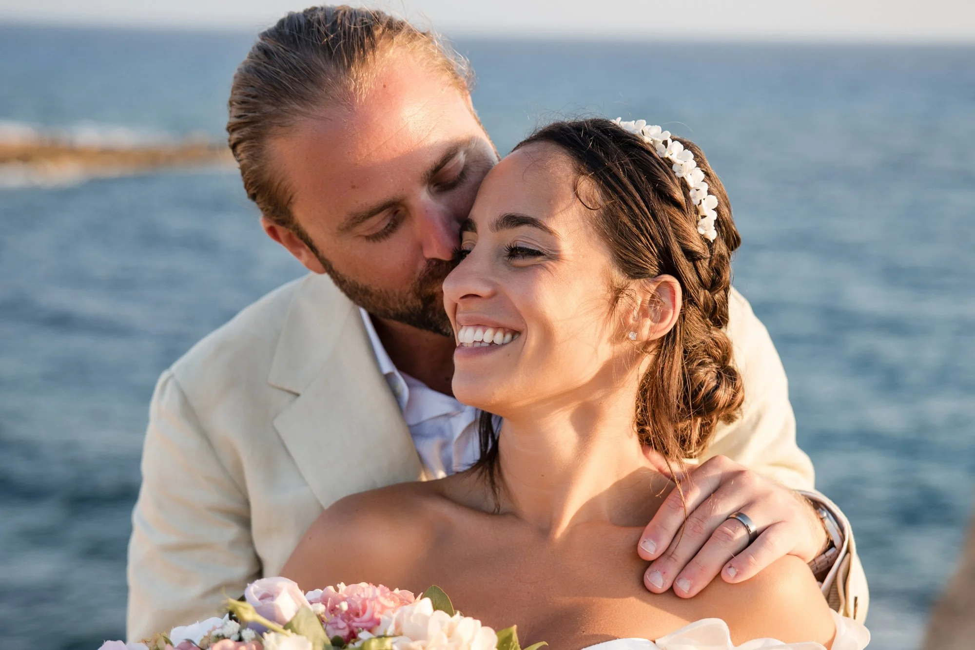 A happy couple embracing outdoors by the water, with the man kissing the woman on her forehead and she smiling, holding a bouquet of flowers.