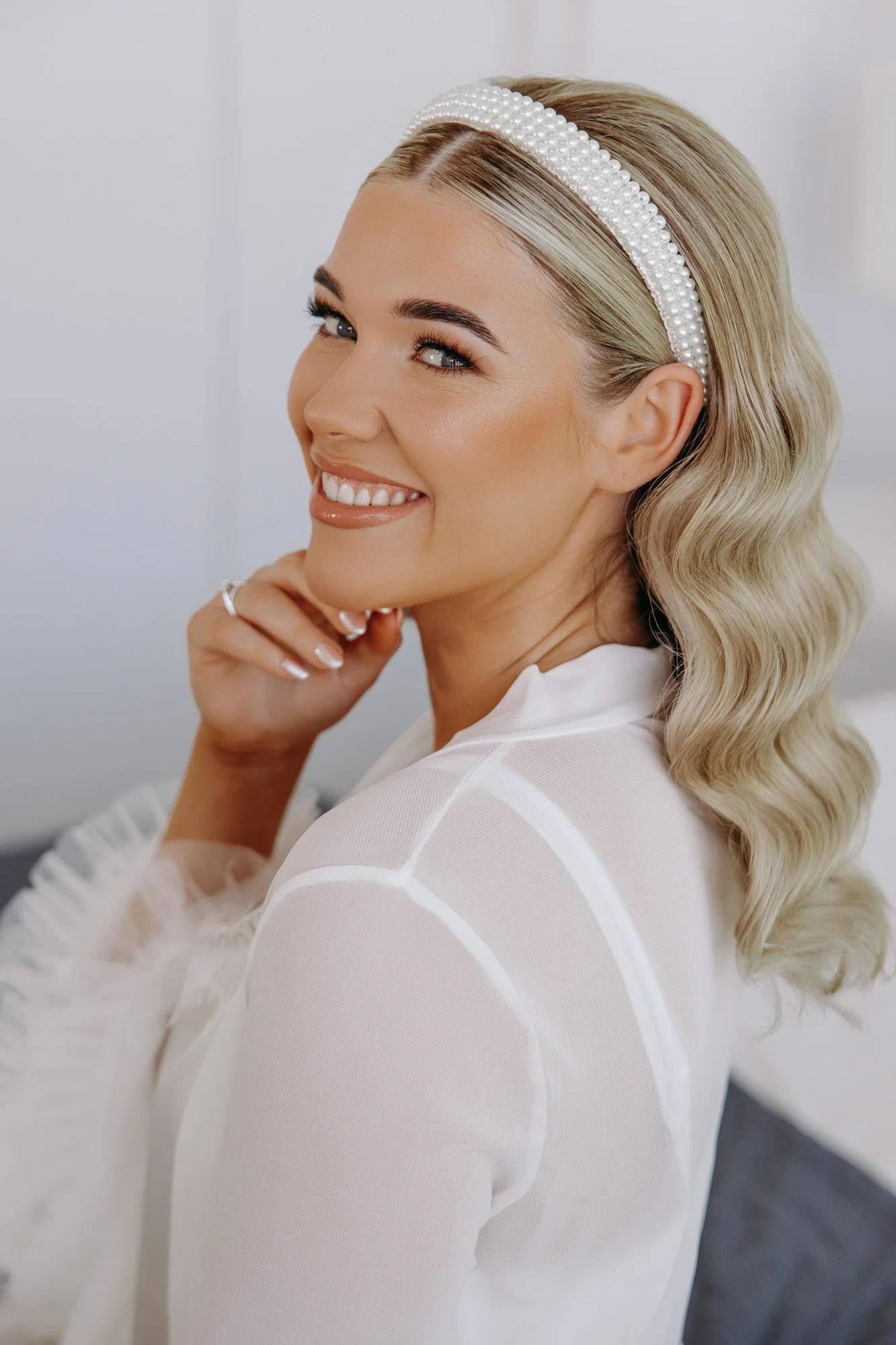 A smiling woman with blonde wavy hair wearing a pearl headband and a white top.