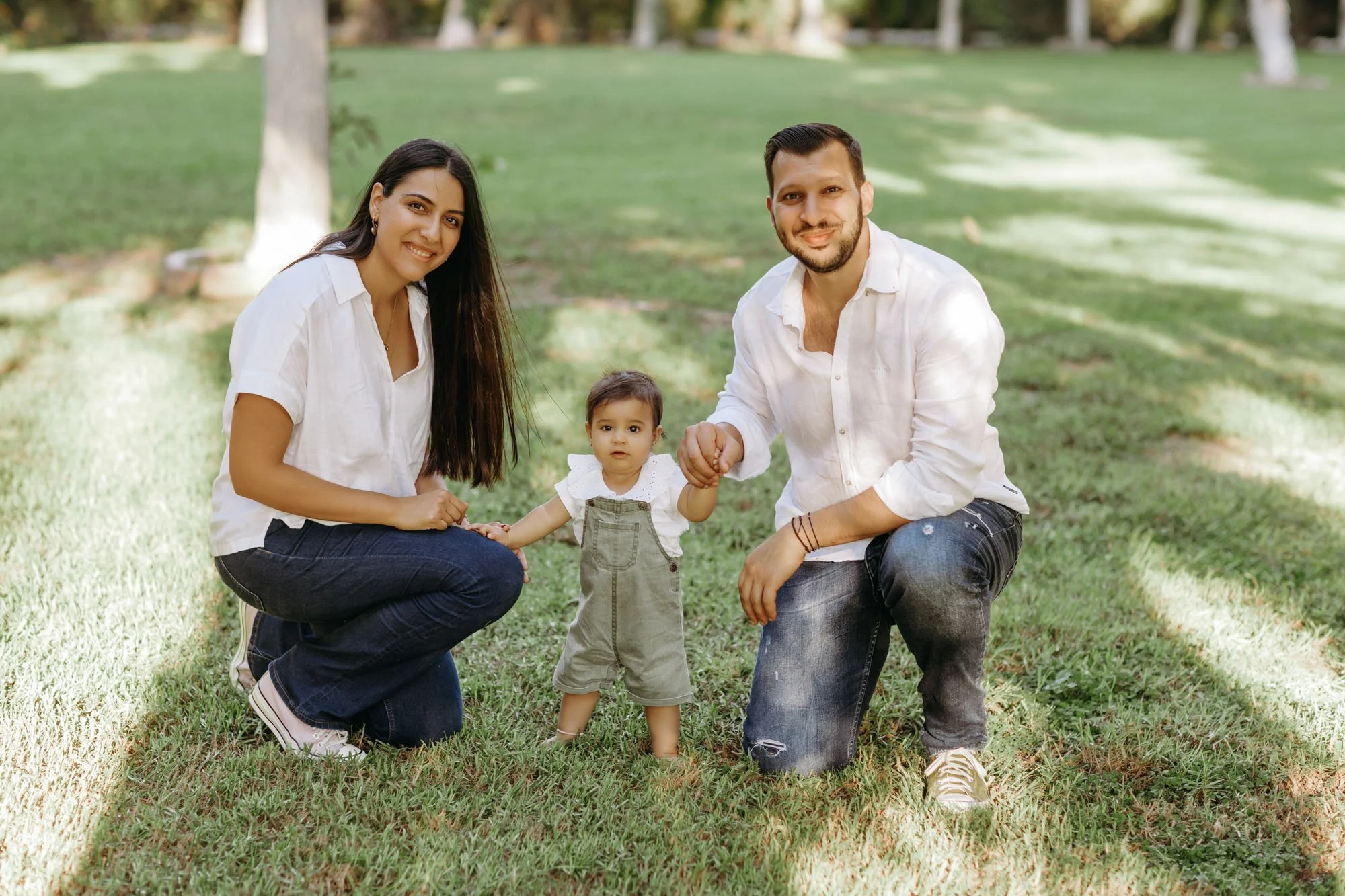 A family of three, a woman, a man, and a toddler, outdoors on a grassy area, holding hands and smiling at the camera.