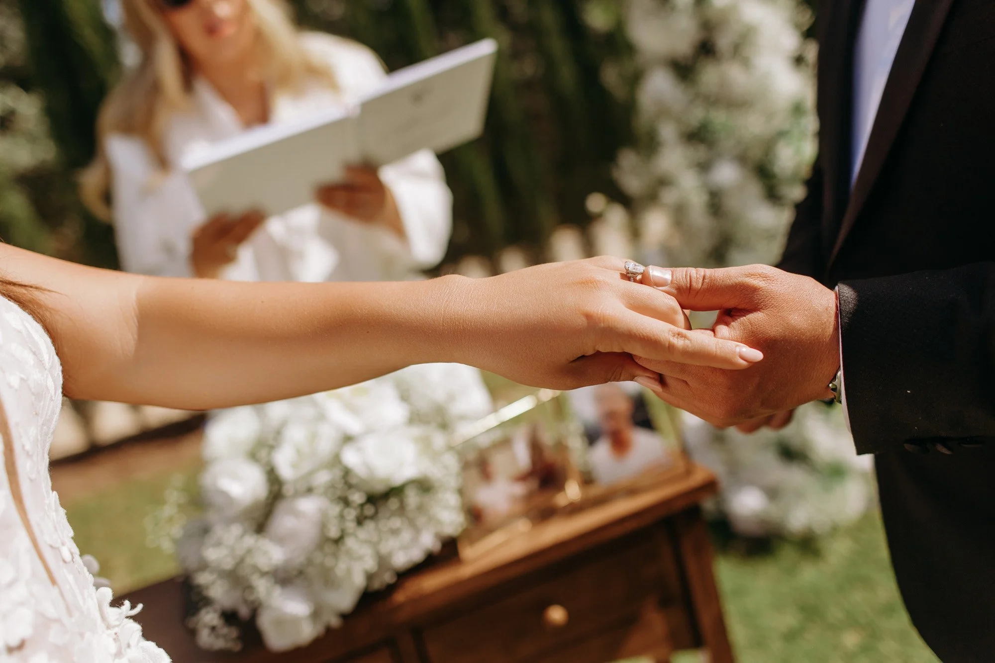 Close-up of a bride and groom holding hands during a wedding ceremony, with a woman reading vows or a speech in the background.