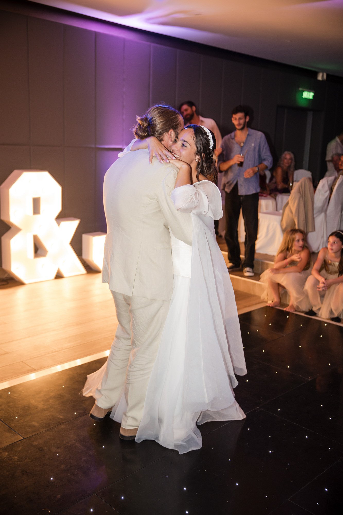 A bride and groom share a dance at their wedding reception, with guests watching and smiling in the background, illuminated by soft lighting and large light-up letters.