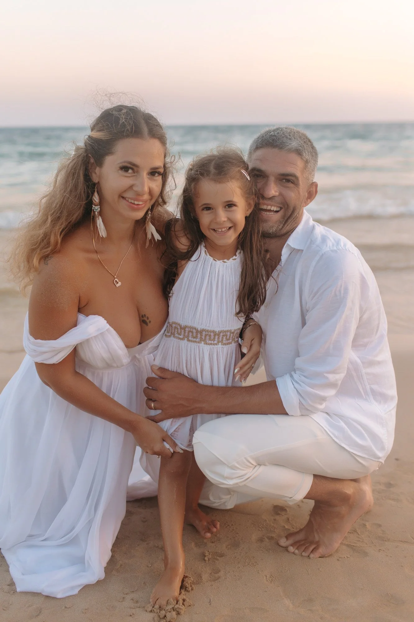 A family of three, including a woman, a man, and a young girl, smiling and posing together on a sandy beach with the ocean and sky in the background at sunset.