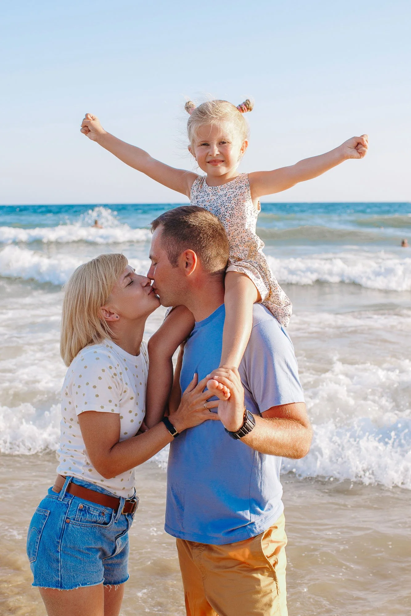 A family at the beach sharing a kiss while a young girl is sitting on her dad's shoulders with arms raised in joy. The family appears to be enjoying a sunny day by the ocean.