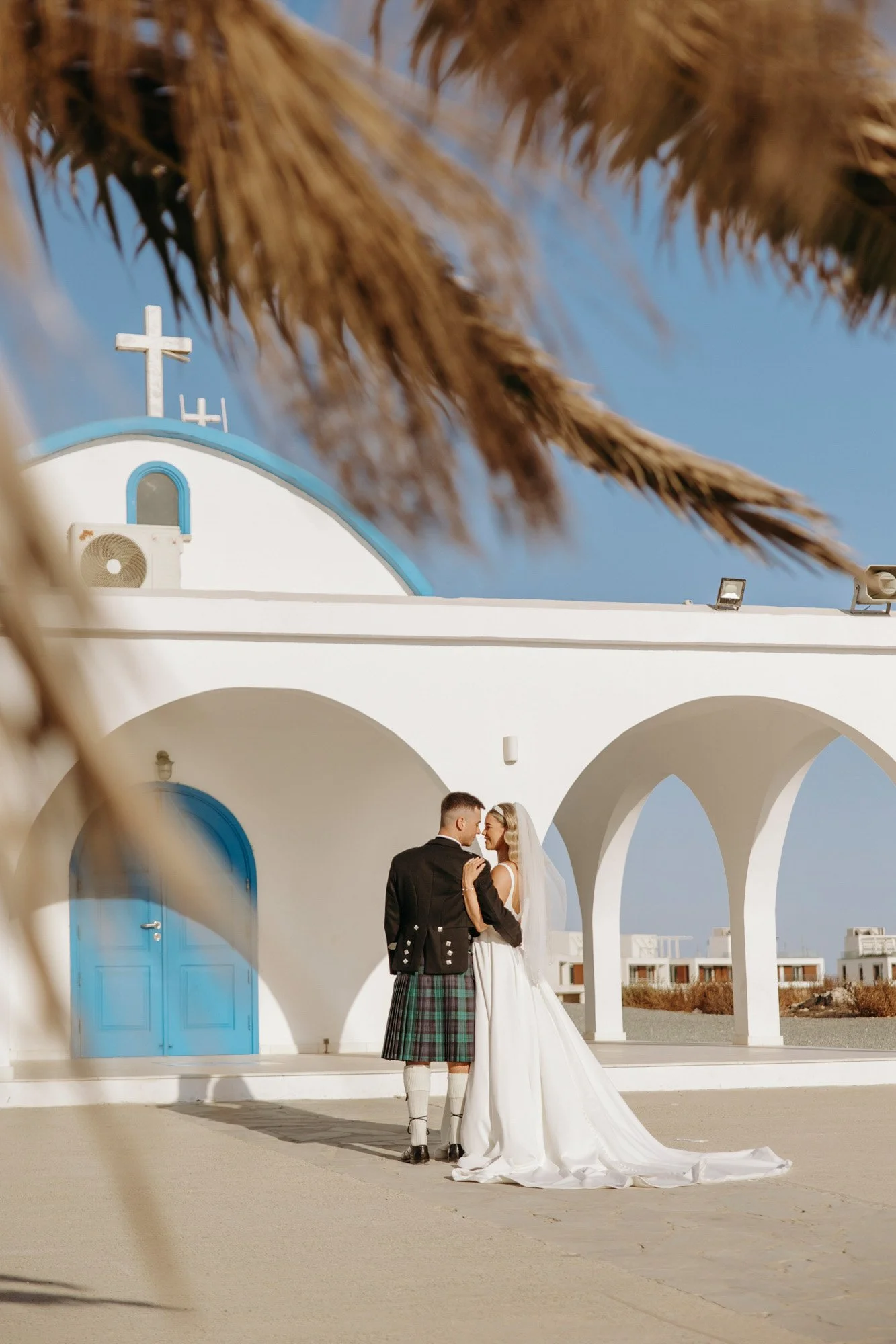 A bride and groom standing close together in front of a white Greek-style chapel with blue accents, under arches with the sky behind.