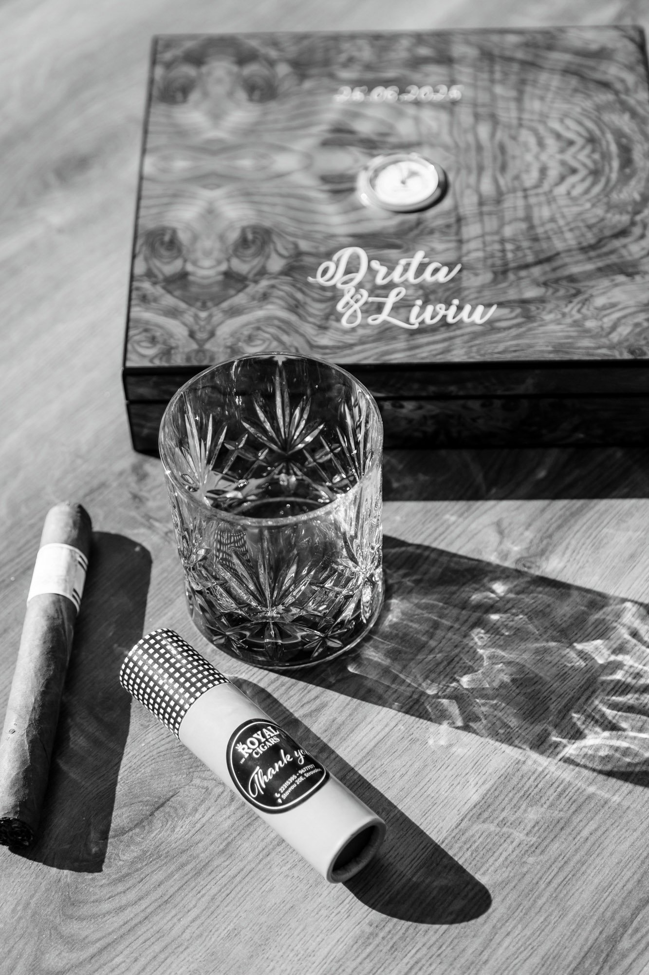 A glass of water, a cigar, and a lighter on a wooden table with a decorative box in the background. The box has a date and the words "Druta & Liviu" written on it.