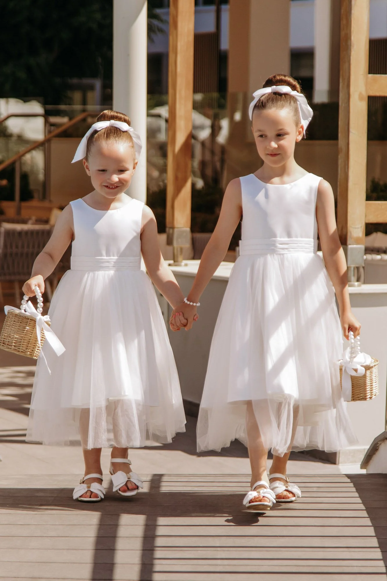 Two young girls in white dresses holding hands, carrying small basket purses, walking outdoors on a wooden deck, dressed for a special occasion like a wedding.