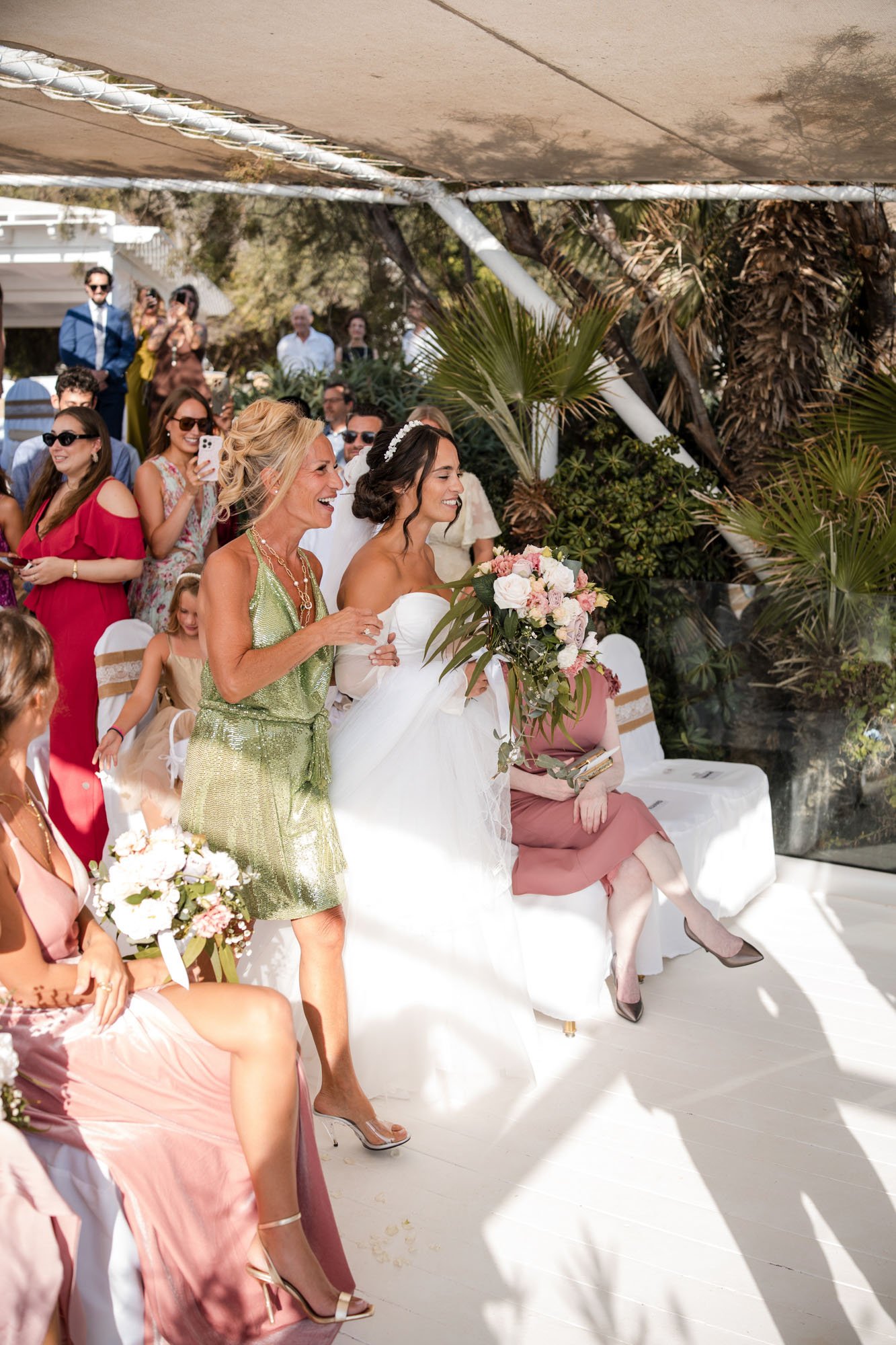 A bride in a white wedding dress holding a bouquet of pink and white flowers, standing next to a smiling woman in a green dress at a wedding ceremony, with guests seated and standing around them, outdoors with greenery and trees in the background.