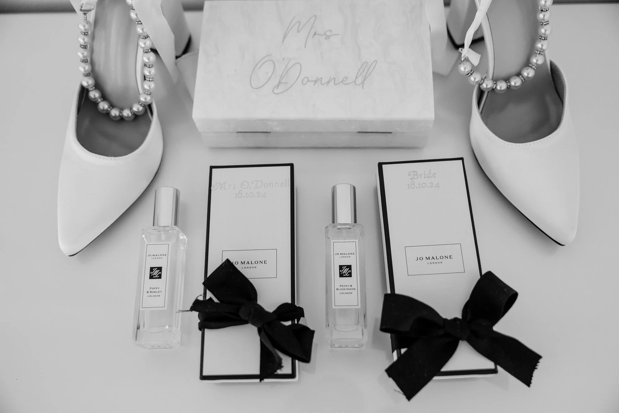 Wedding accessories including white shoes with pearl ankle straps, a bouquet of white flowers, perfume bottles, and personalized name cards for the bride and groom.