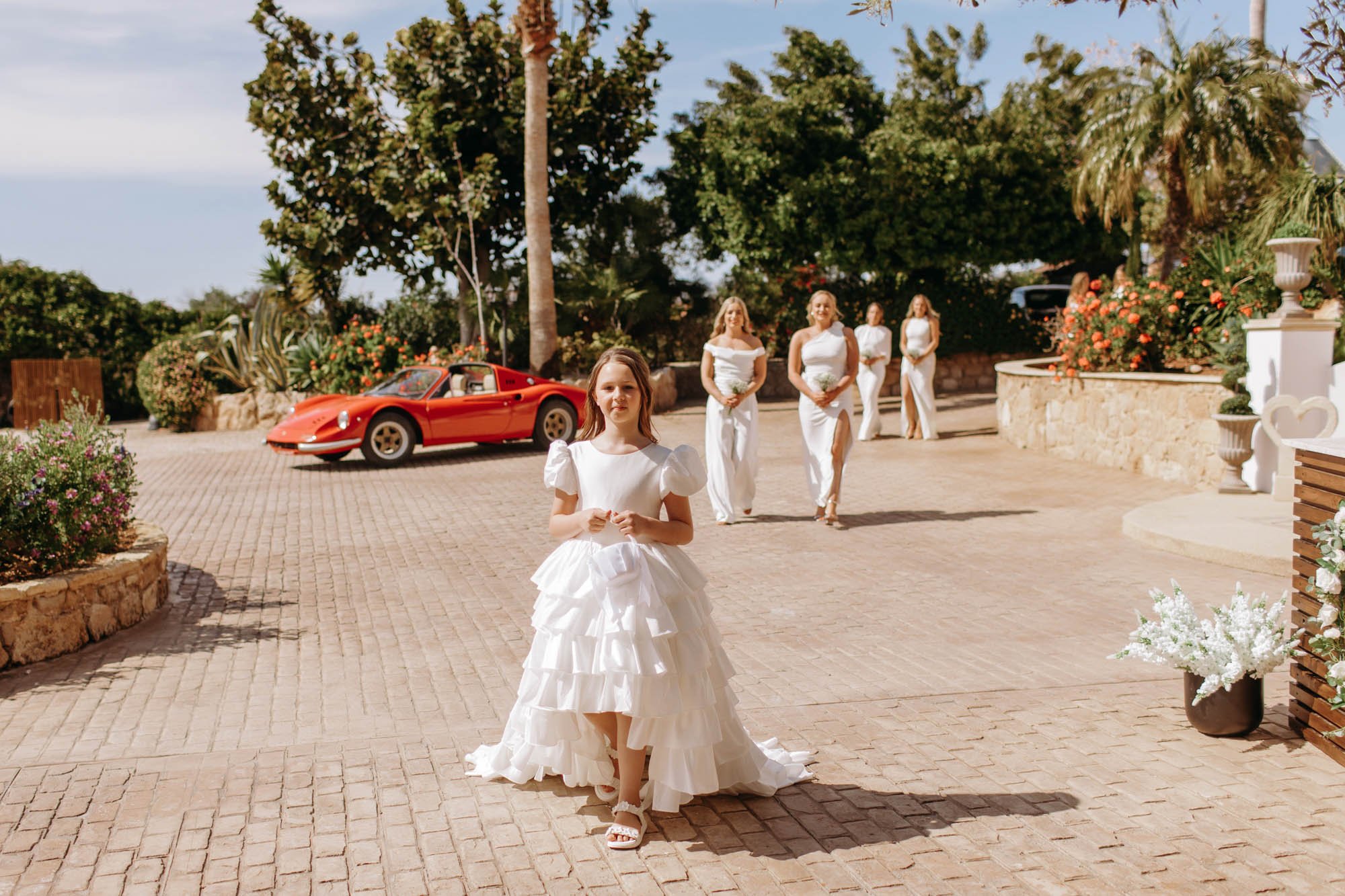 A young girl in a white ruffled dress walking towards camera, with four women in white dresses walking behind her, outdoors in a landscaped area with flowers, trees, and a vintage red sports car.