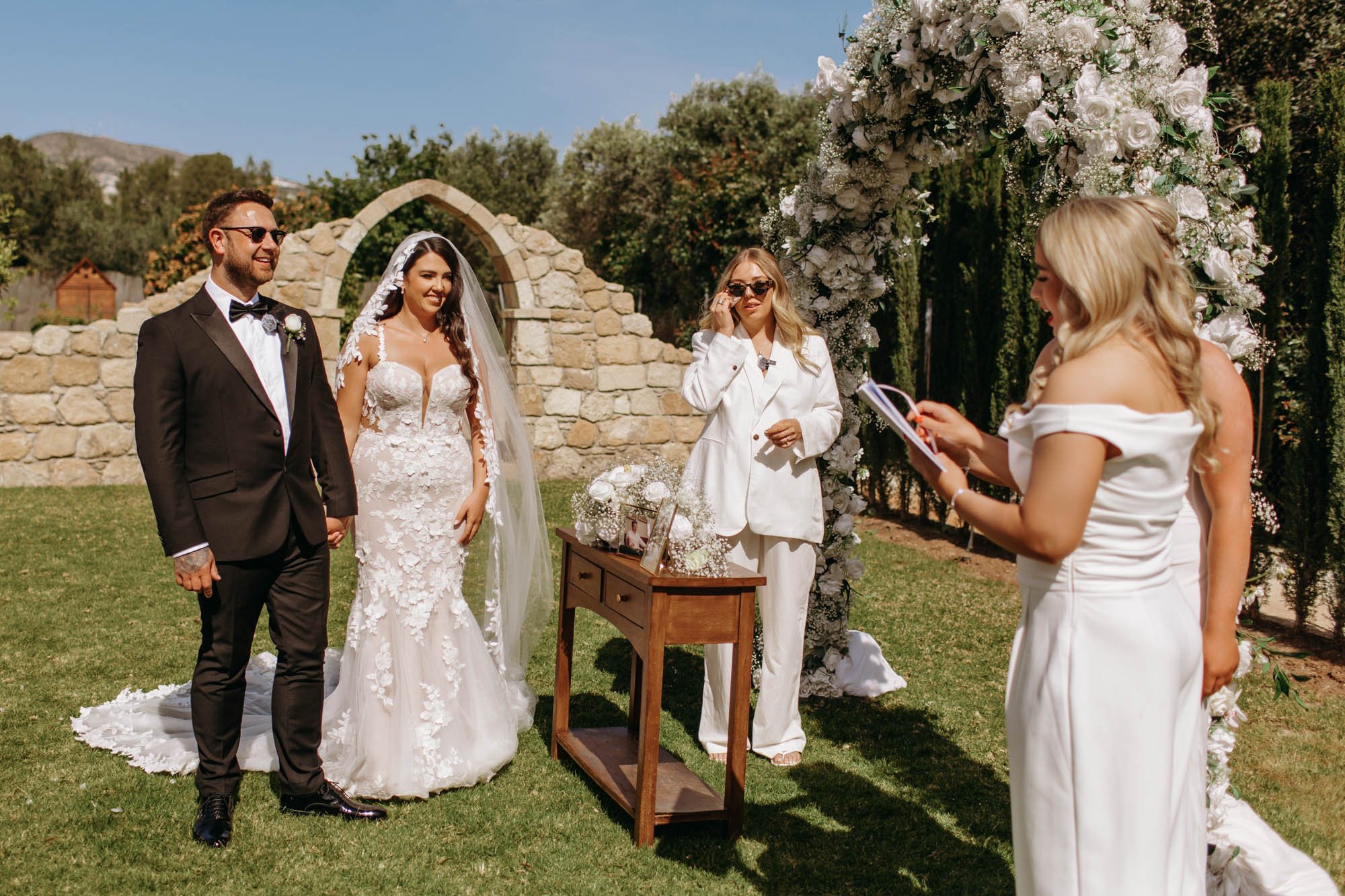 A wedding ceremony outdoors with the bride and groom standing together, the bride wearing a lace wedding dress and ponytail, the groom in a tuxedo with sunglasses. Two women in white dresses officiate, one holding a script and the other touching her 