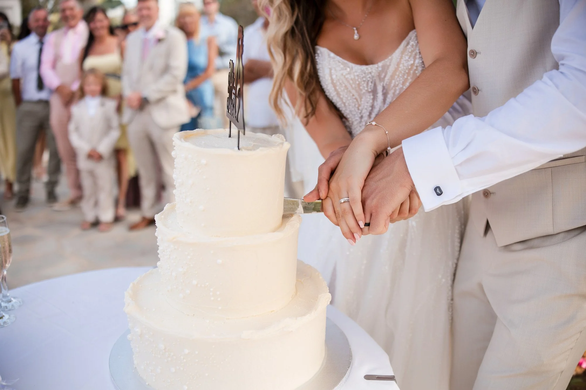 A bride and groom cutting a three-tiered wedding cake together during their wedding celebration, with guests in the background.