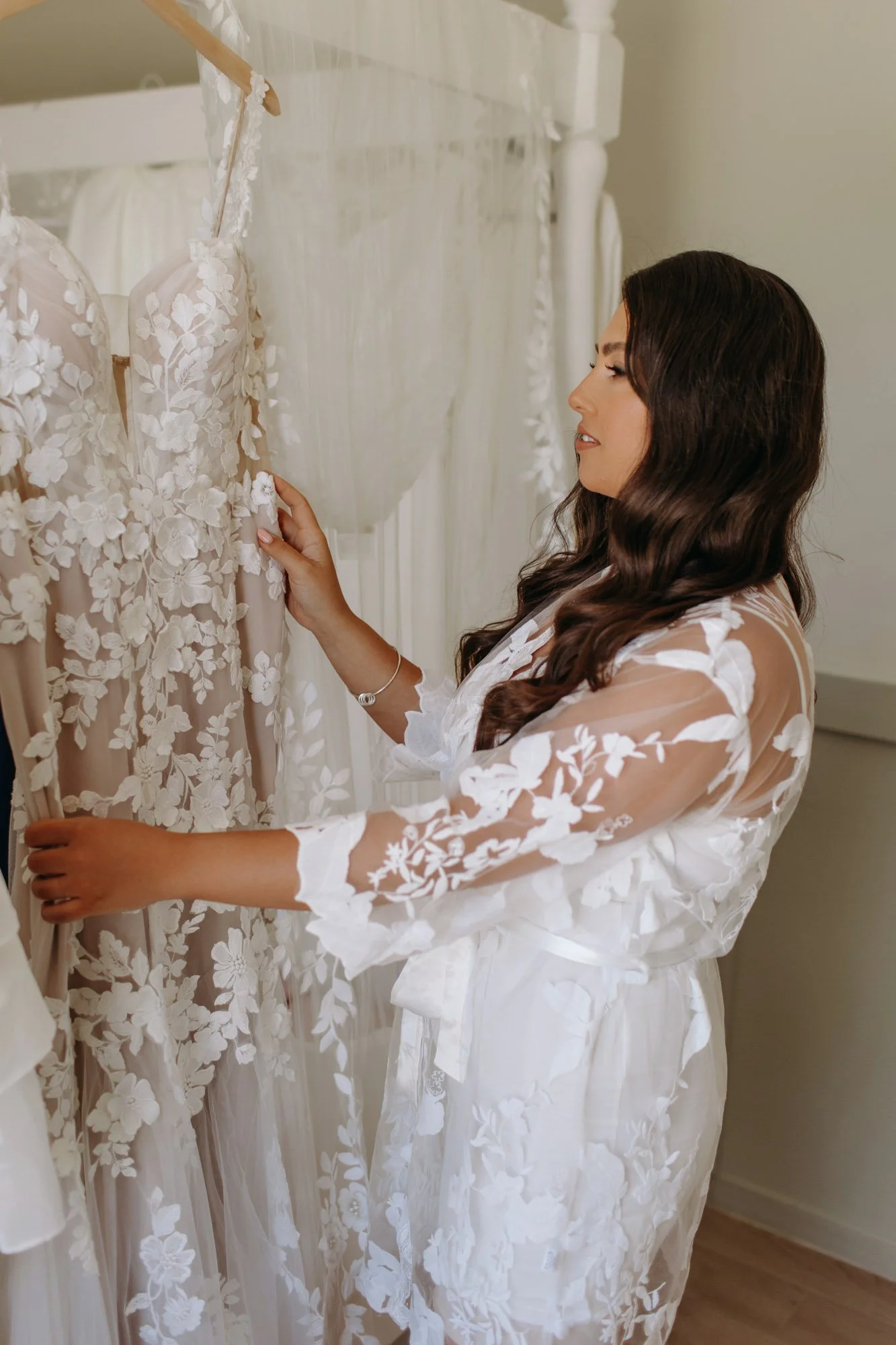 A woman in a sheer, white floral robe looks at a wedding dress hanging on a rack.