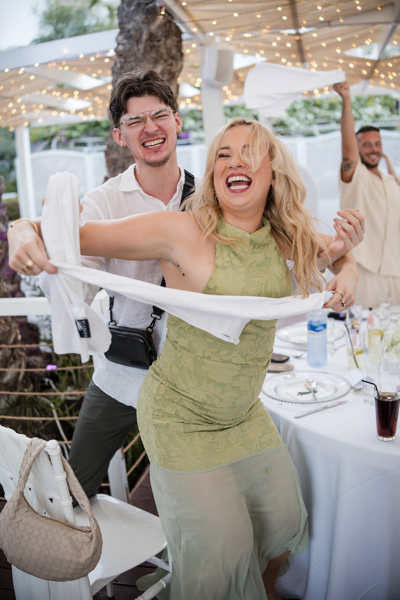 People celebrating at an outdoor event, with a woman in a green dress holding a white cloth and smiling, and a man behind her with a joyful expression, under string lights.