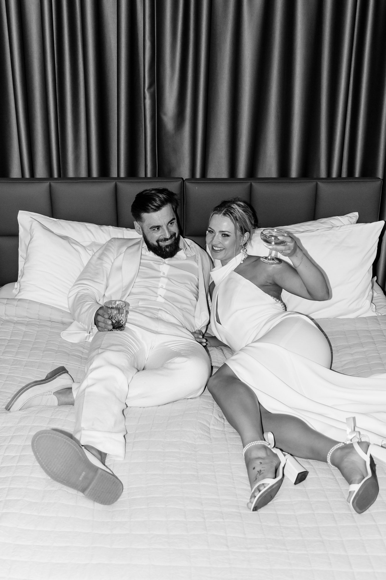 A couple in formal attire lying on a bed, smiling, holding drinks, with a dark curtain in the background.