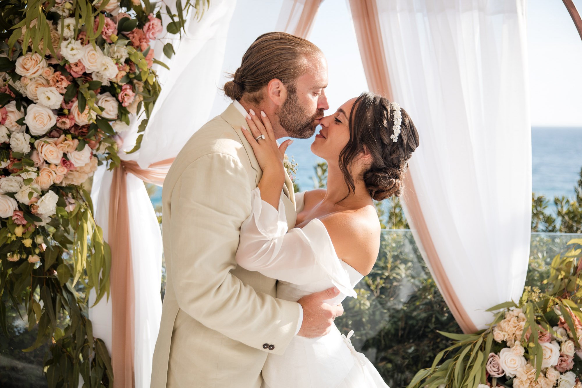 A bride and groom sharing a kiss during their wedding ceremony outdoors with ocean in background, surrounded by floral decorations and sheer curtains.