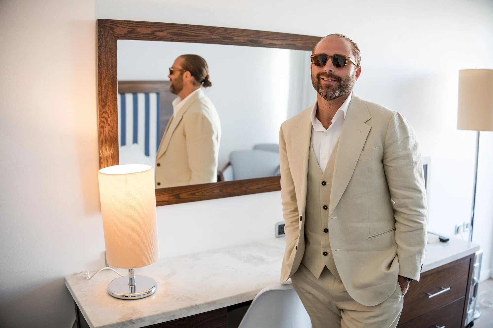 Man with beard and sunglasses in a beige suit standing in a hotel room next to a dresser with a lamp, reflected in a large mirror behind him.