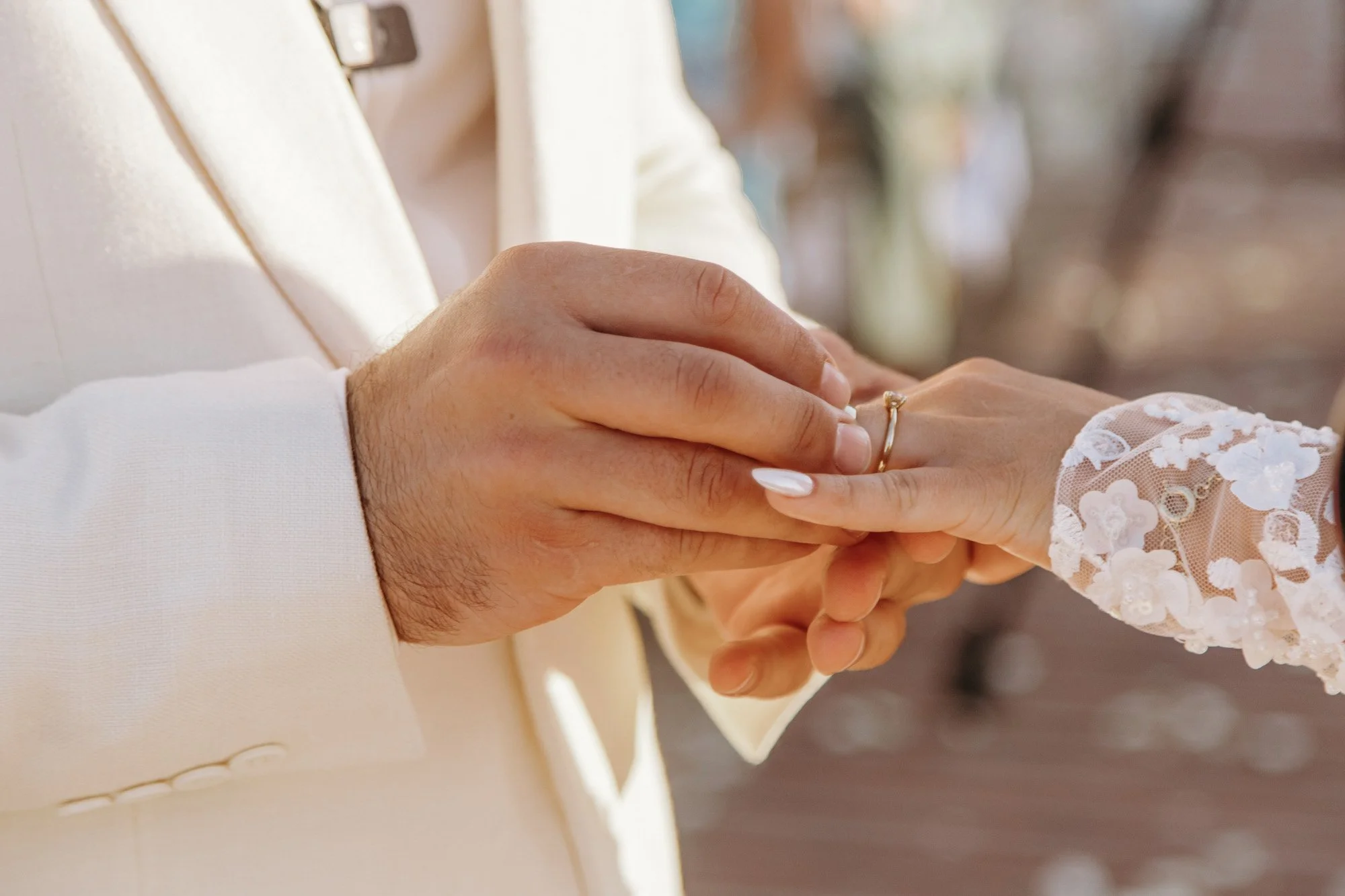 Person in a white suit holding a woman's hand, placing a ring on her finger, with lace sleeve detail visible.
