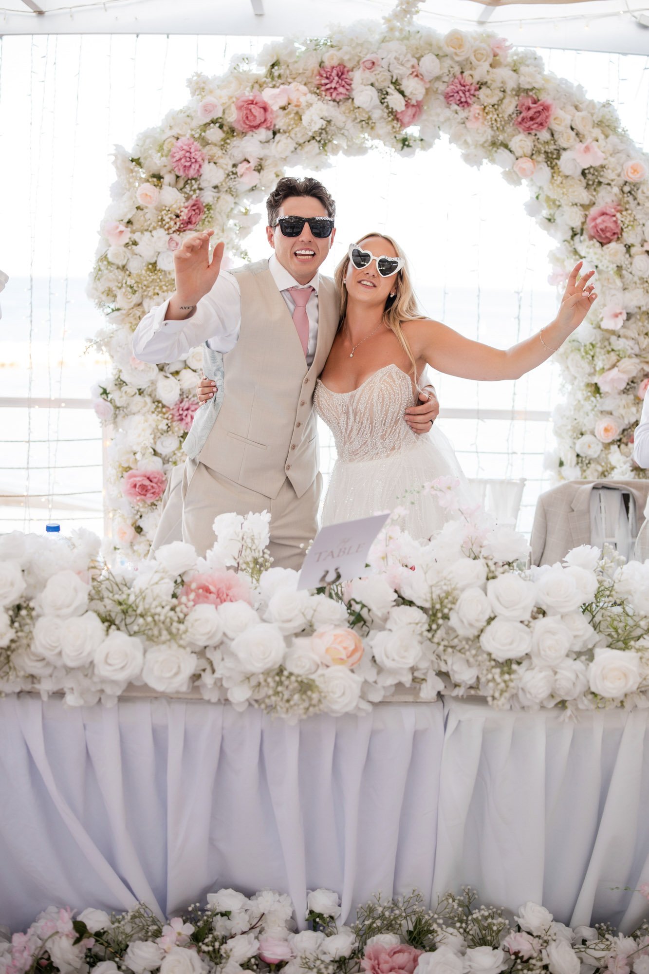 A bride and groom dancing at their wedding reception, wearing sunglasses, standing in front of a large floral arch with pink and white flowers, surrounded by an abundance of white flowers on a decorated table.