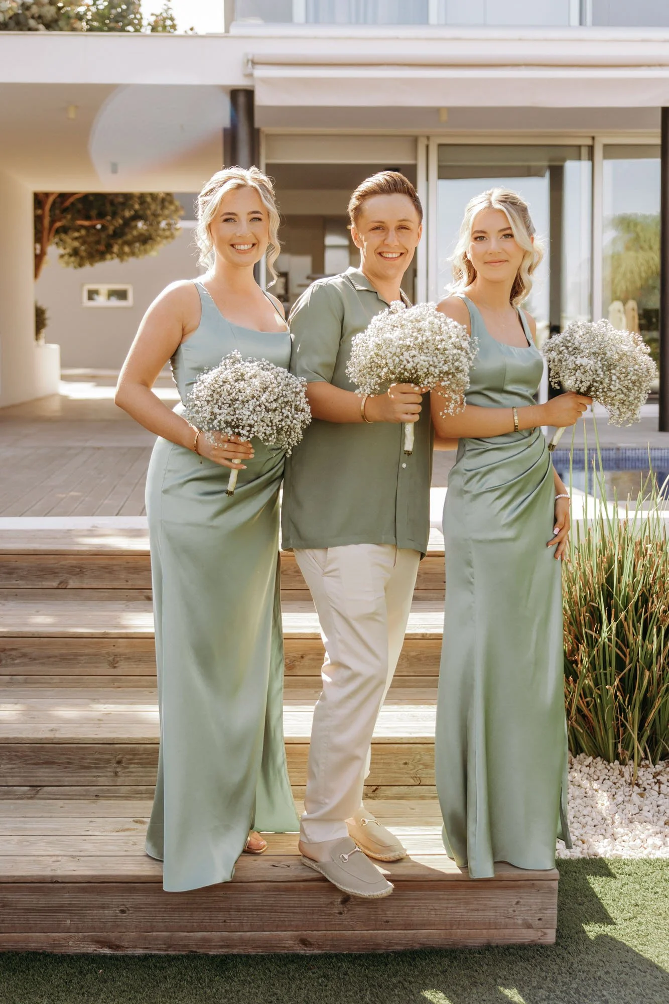 Three women and one man standing on a wooden deck outdoors, each holding a bouquet of white flowers, dressed in pastel-colored clothing, smiling at the camera.