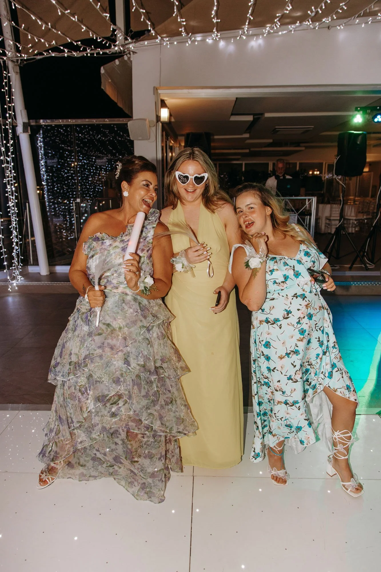 Three women dressed in colorful dresses enjoying a party, with decorative string lights hanging from the ceiling behind them.