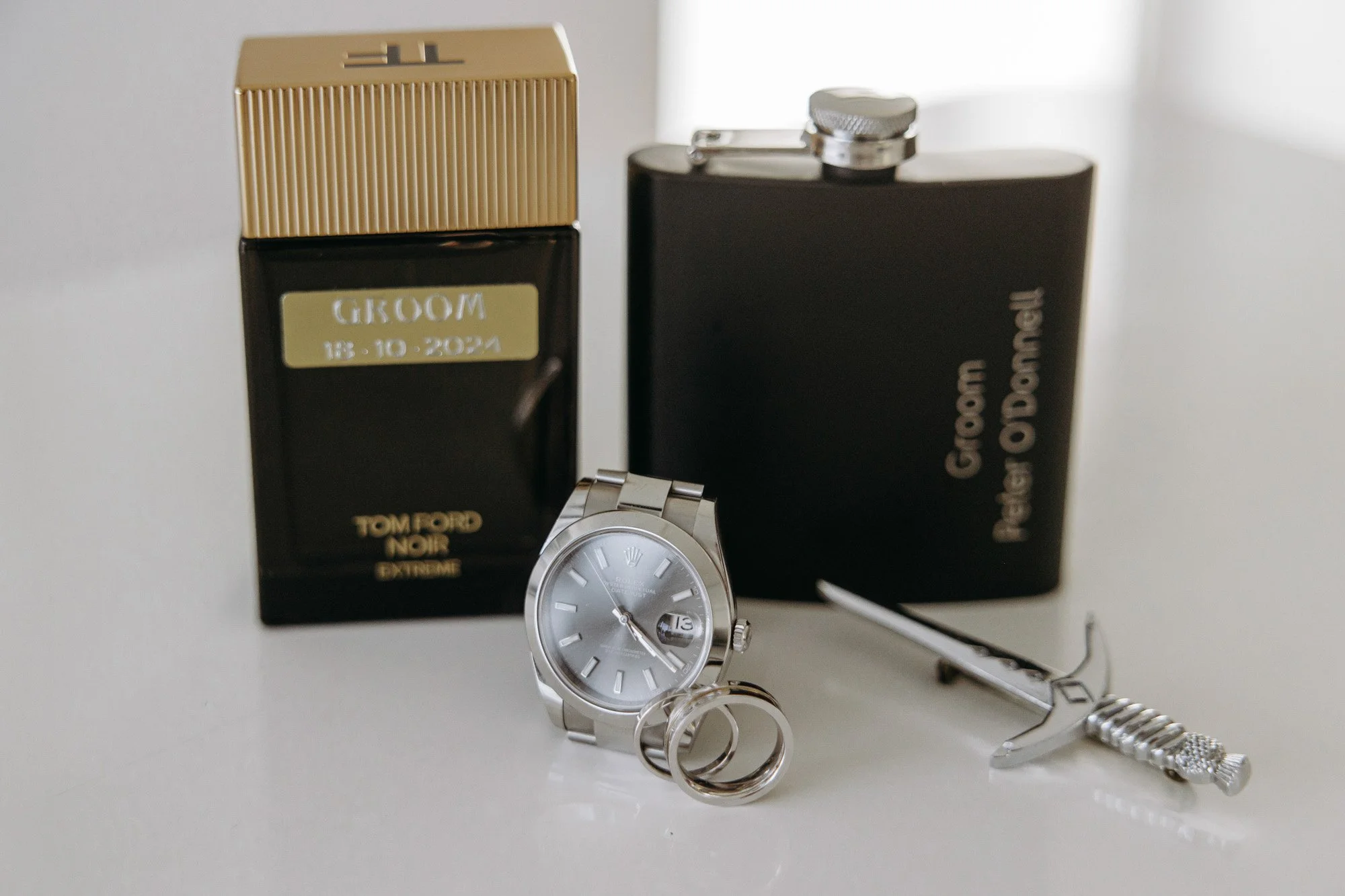 Tom Ford Noir Extreme perfume bottles, a silver wristwatch, two wedding rings, and a small sword-shaped dagger with a jeweled handle on a light-colored surface.