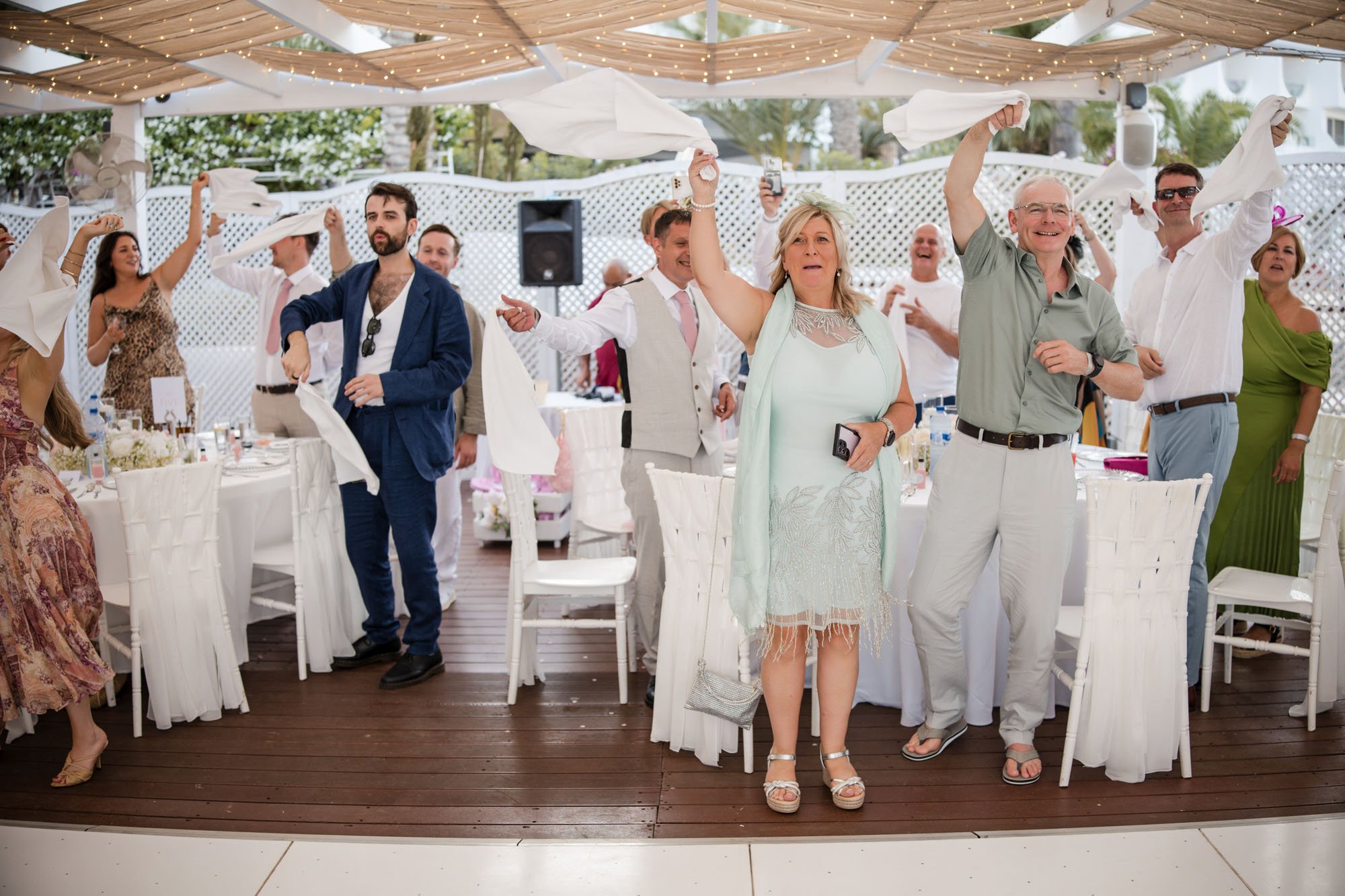 Guests dancing under a decorated outdoor canopy at a celebration or wedding reception, with white chairs, tables, and string lights.