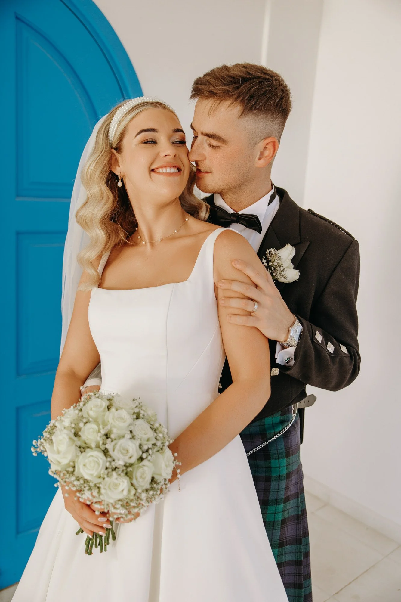 A bride and groom sharing a joyful moment on their wedding day. The bride wears a white dress, holding a bouquet of white roses, with blonde wavy hair and a pearl headband. The groom is dressed in a black uniform with a tartan kilt, black bow tie, an