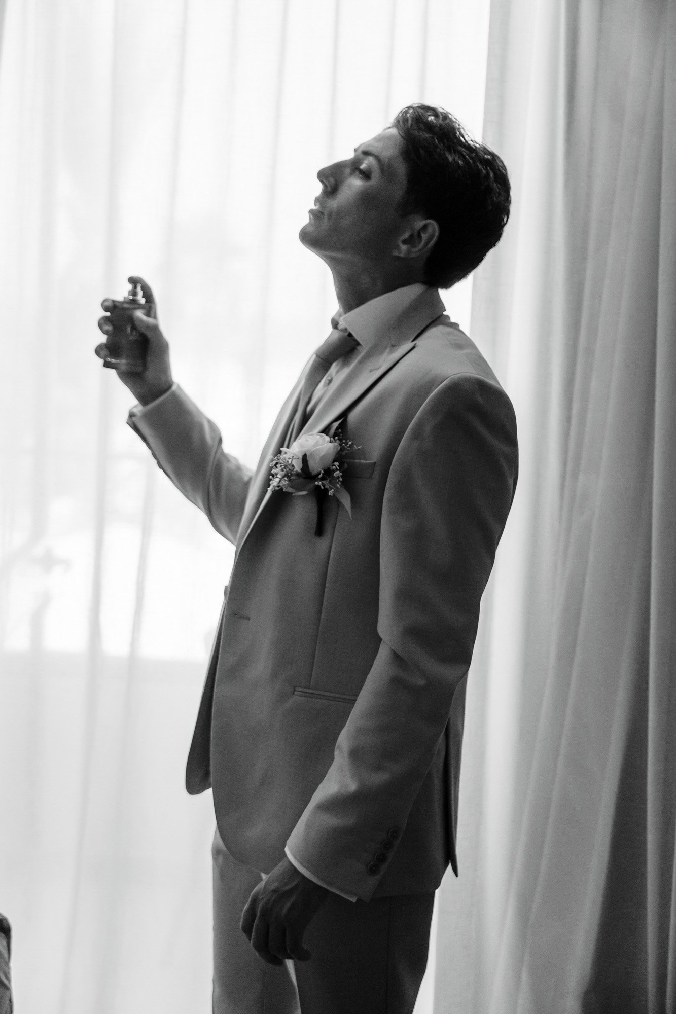 A man in a suit preparing to spray cologne, standing in front of curtains. The photo is in black and white.