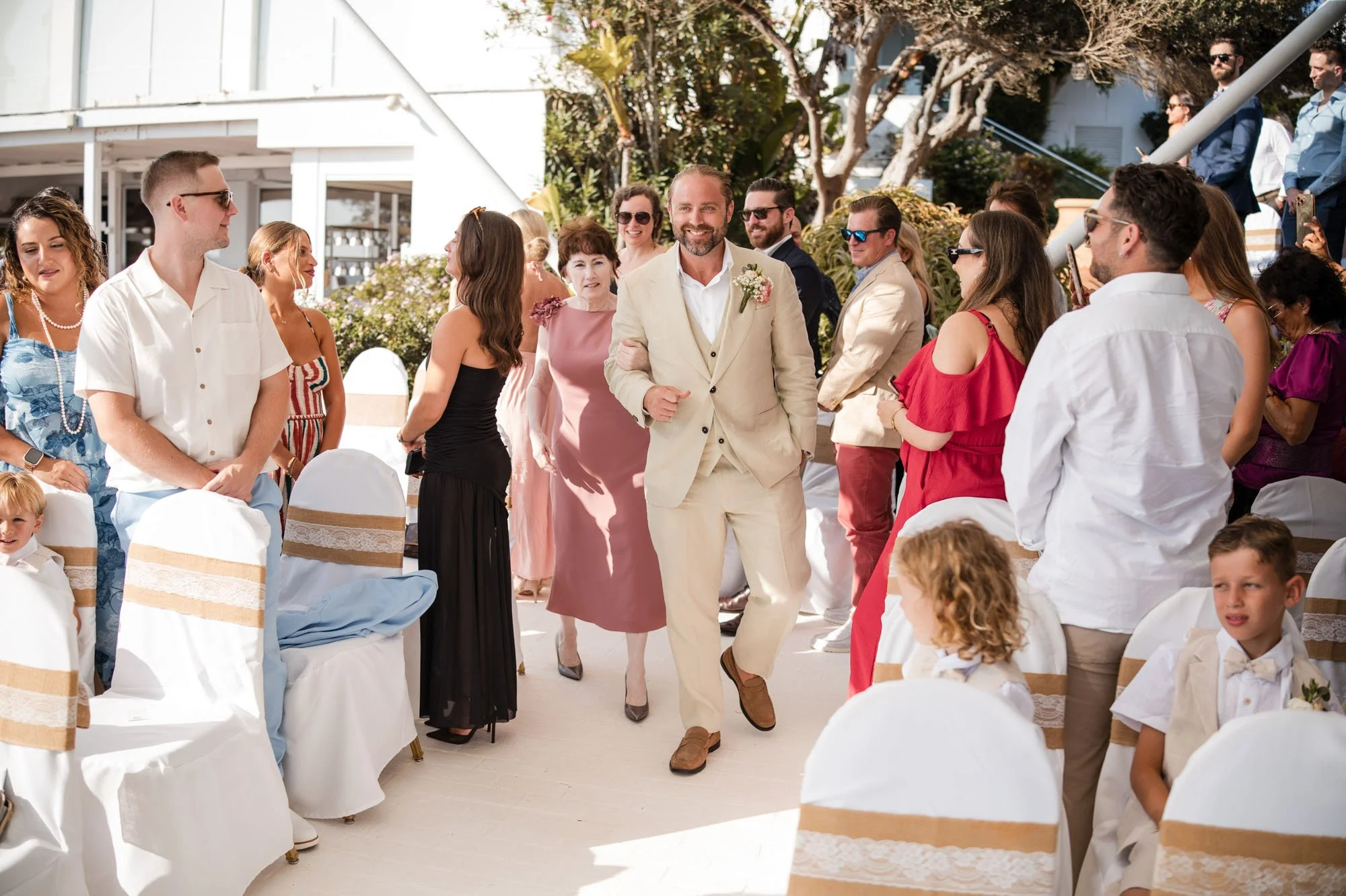 Man in beige suit walking down aisle at outdoor wedding ceremony, smiling, surrounded by seated and standing guests, with some dressed in colorful attire, and floral decorations in the background.